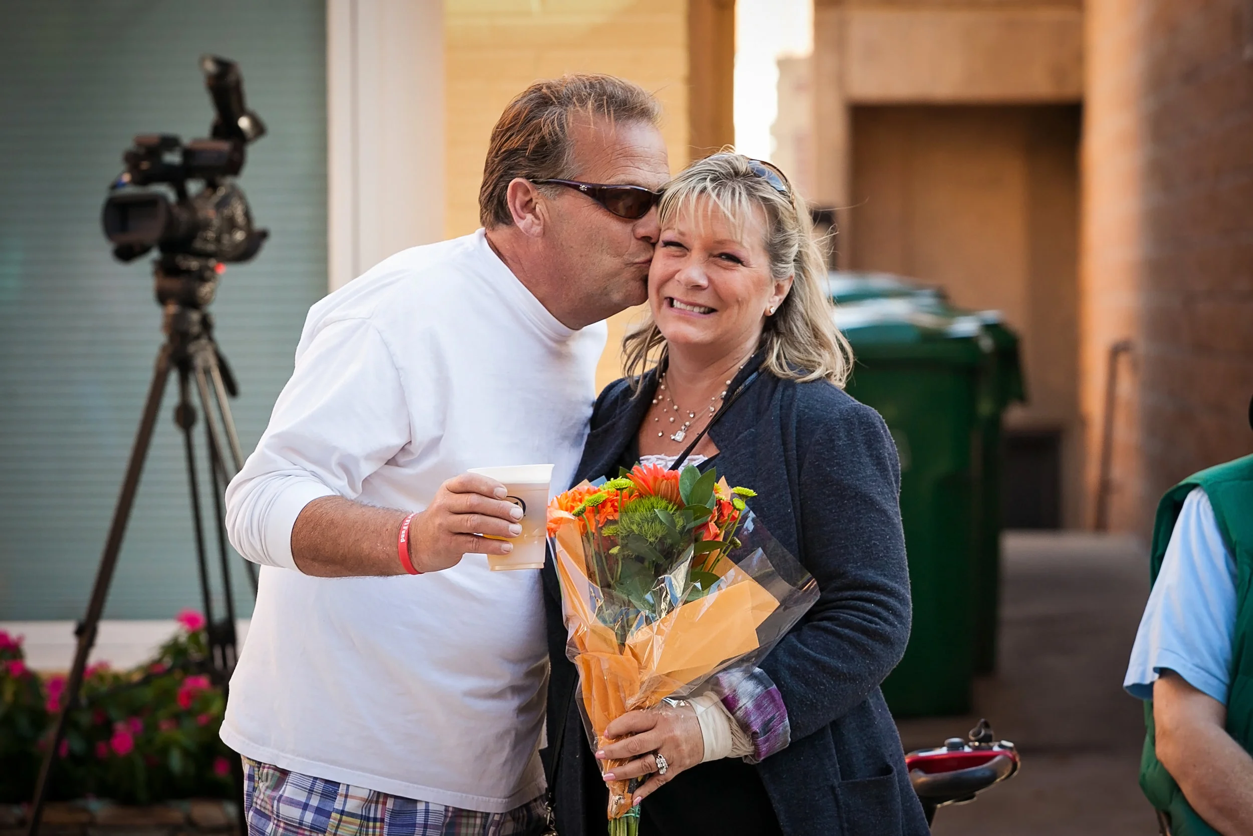 A man kisses a woman on the cheek while holding a beer, and she holds a bouquet of flowers, smiling. They are outdoors with a microphone and flowers in the background.