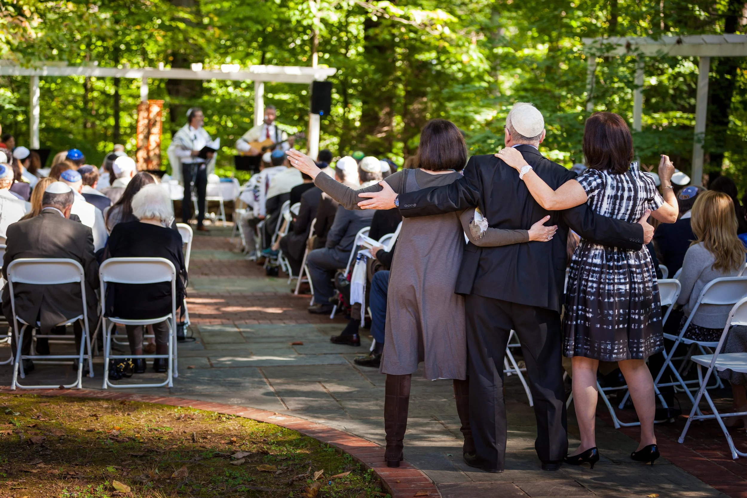 Outdoor gathering with people seated on white chairs, watching musicians performing on stage in a wooded area, with three people in the foreground embracing and celebrating.