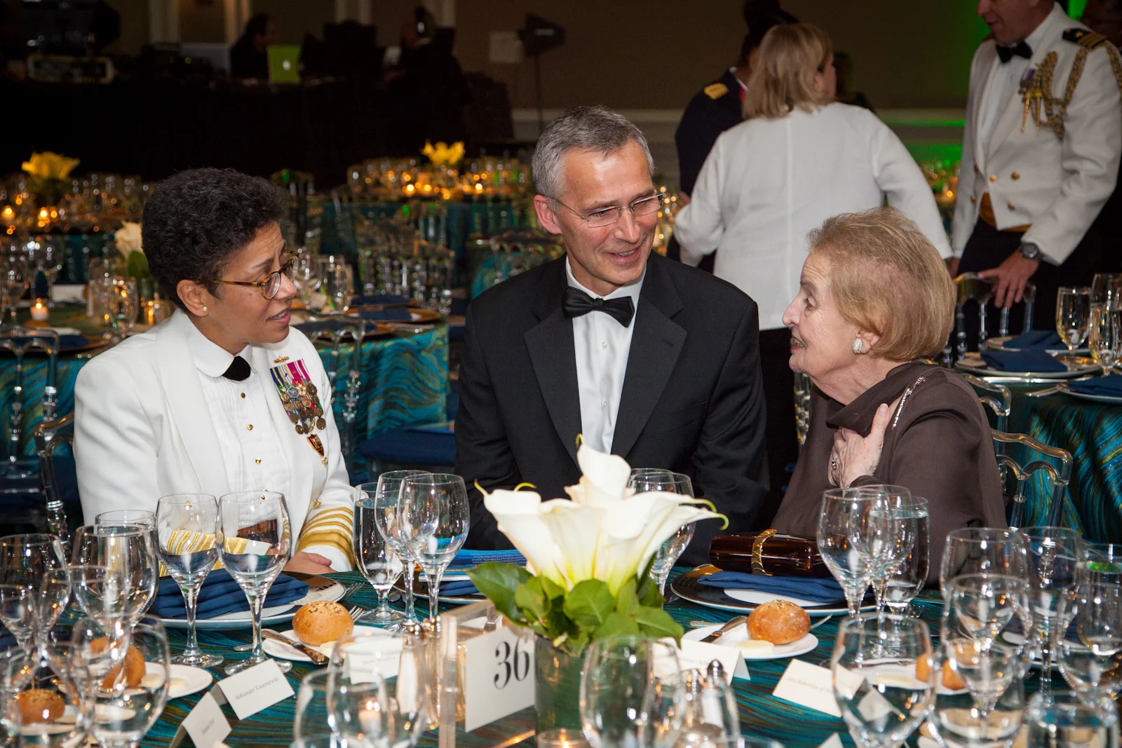 Three people seated at a formal event, engaged in conversation, with decorated tables, floral centerpieces, and uniformed servers in the background.