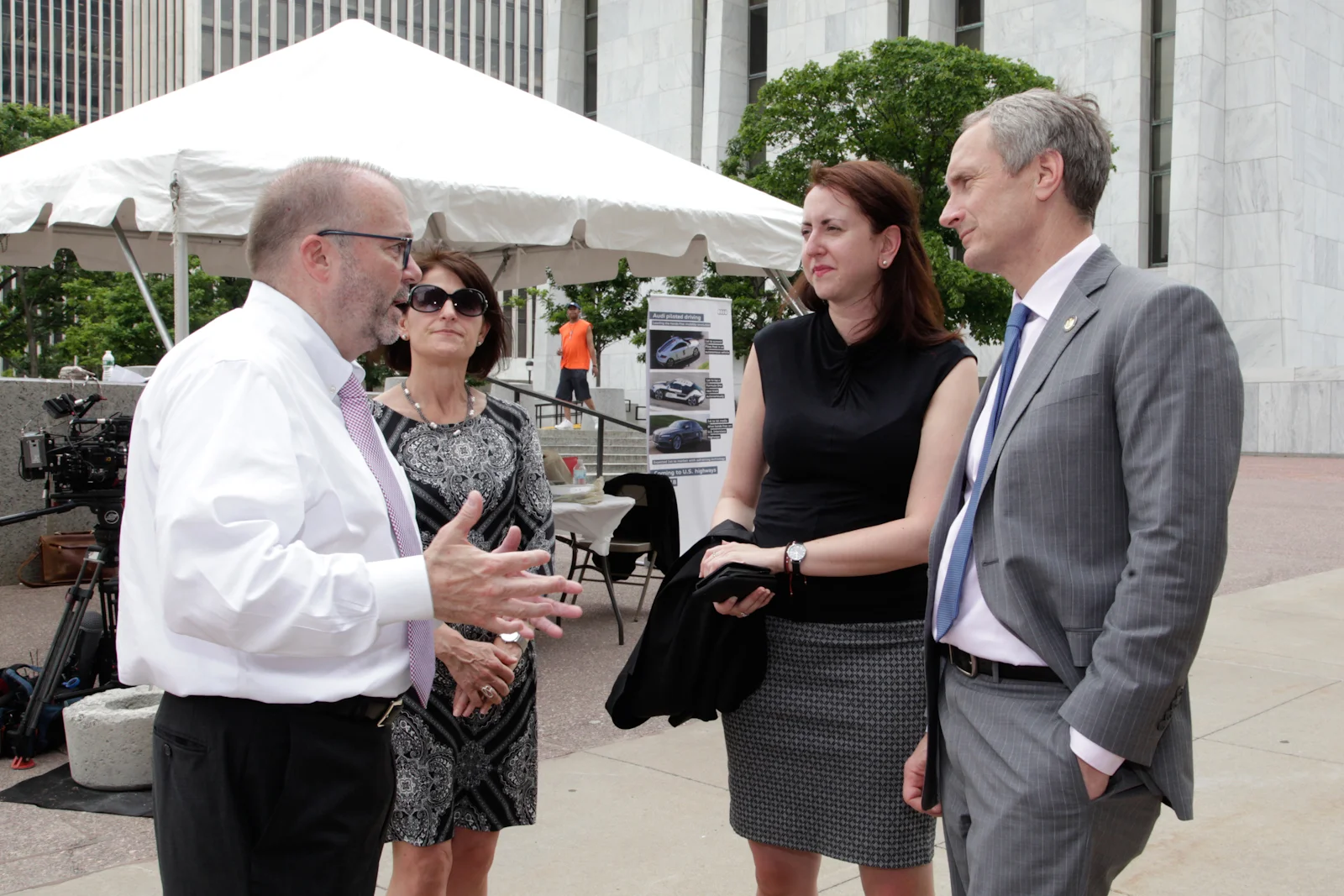 Four adults, two men and two women, engaged in conversation outdoors in front of a white tent, with buildings and trees in the background.