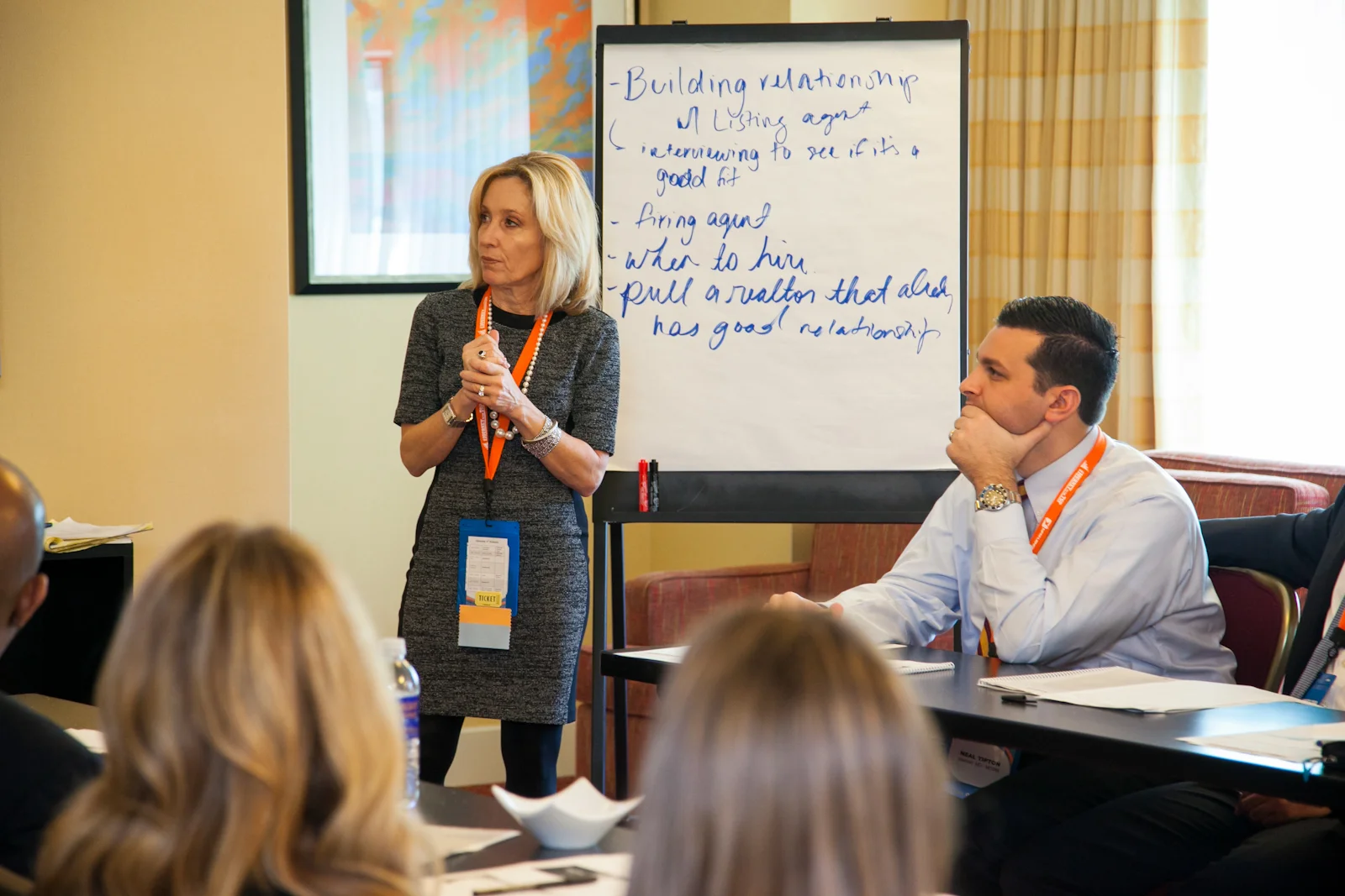 A woman in a gray dress is standing and speaking to a group during a presentation, with a whiteboard behind her with handwritten notes. A man in a white shirt and orange lanyard is sitting at the table, listening attentively.