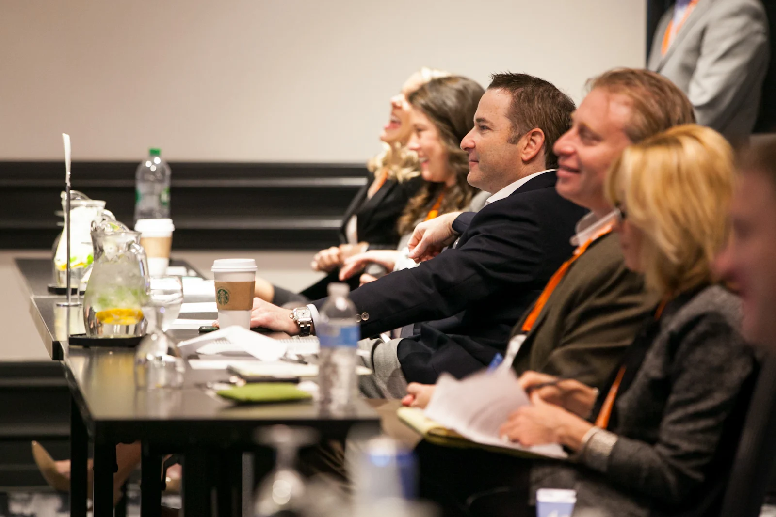 People attending a conference, sitting at a long table with water pitchers, coffee cups, and notebooks, smiling and listening.