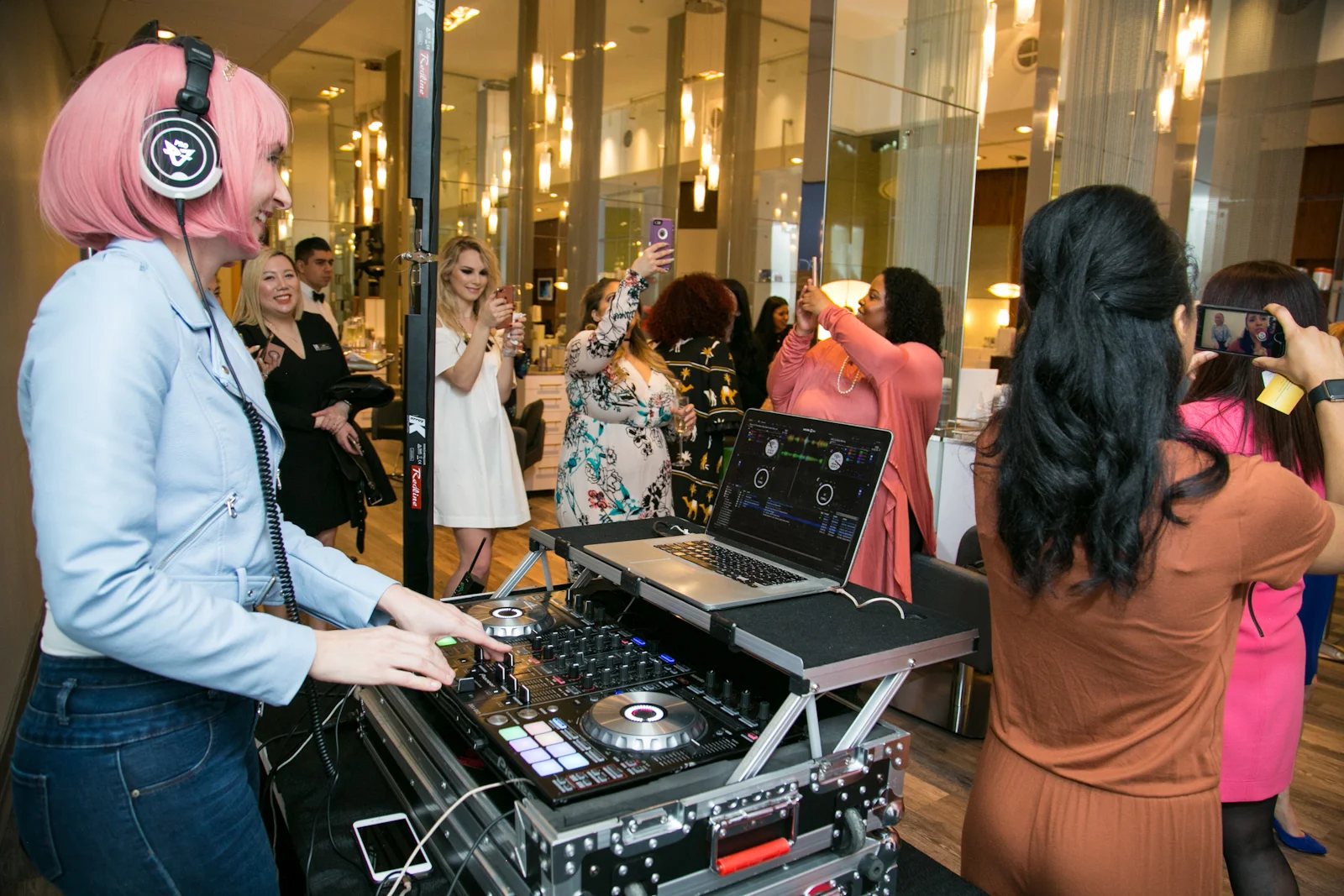 A female DJ with pink hair is performing at a lively indoor event, surrounded by women taking photos and recording videos with their smartphones. The DJ is using professional DJ equipment with a laptop nearby.