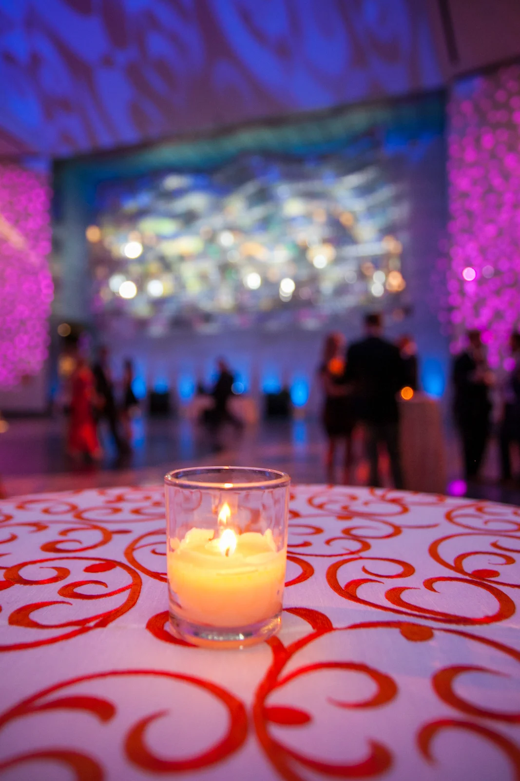 A lit candle in a glass holder on a table with a red and white swirl pattern, in a blurred background with purple and blue lighting and people in tuxedos at a formal event or party.