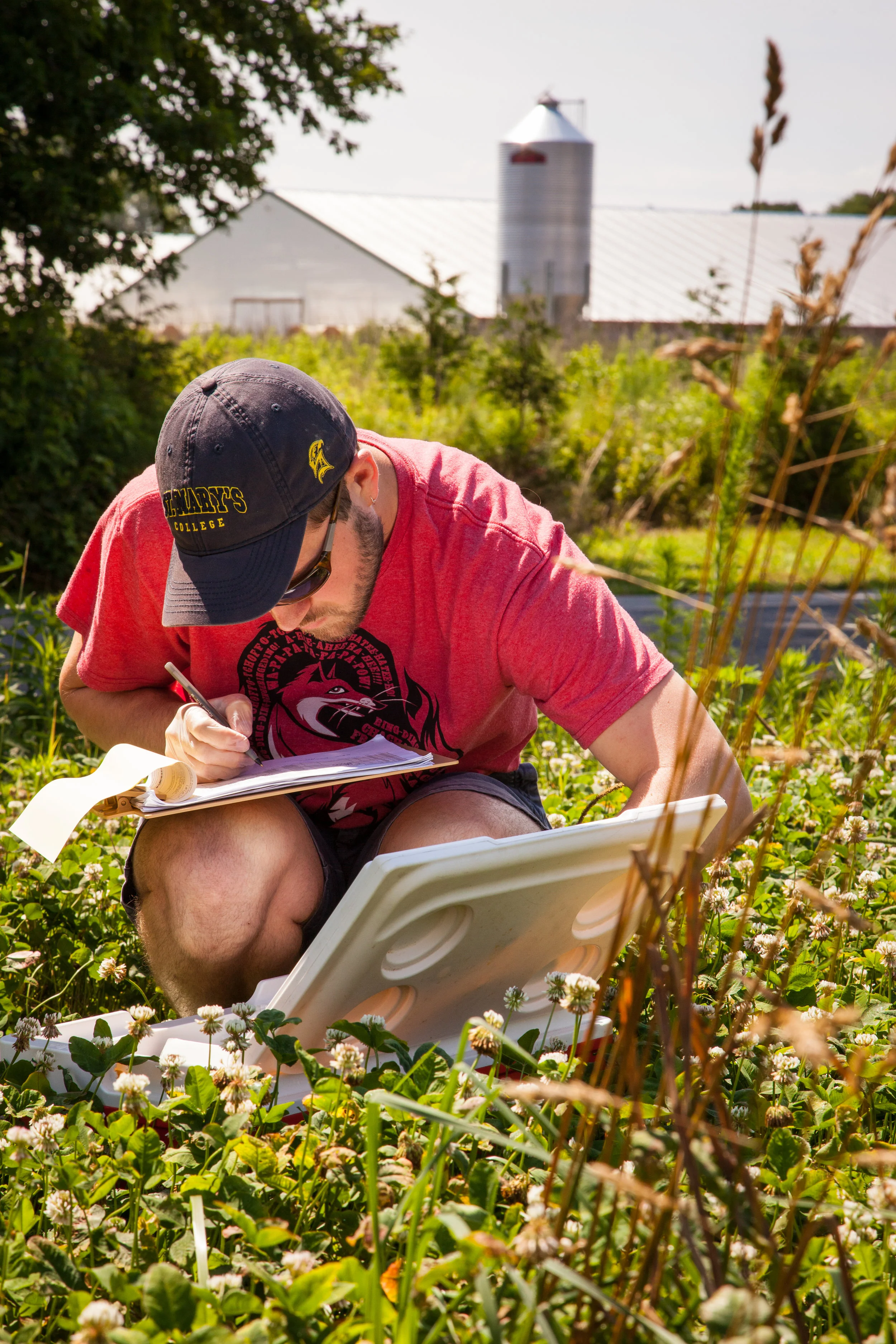 A person wearing a black baseball cap, sunglasses, a red t-shirt, and black shorts kneels in a field of greenery, taking notes with a clipboard, with a large farm grain silo and barn in the background.