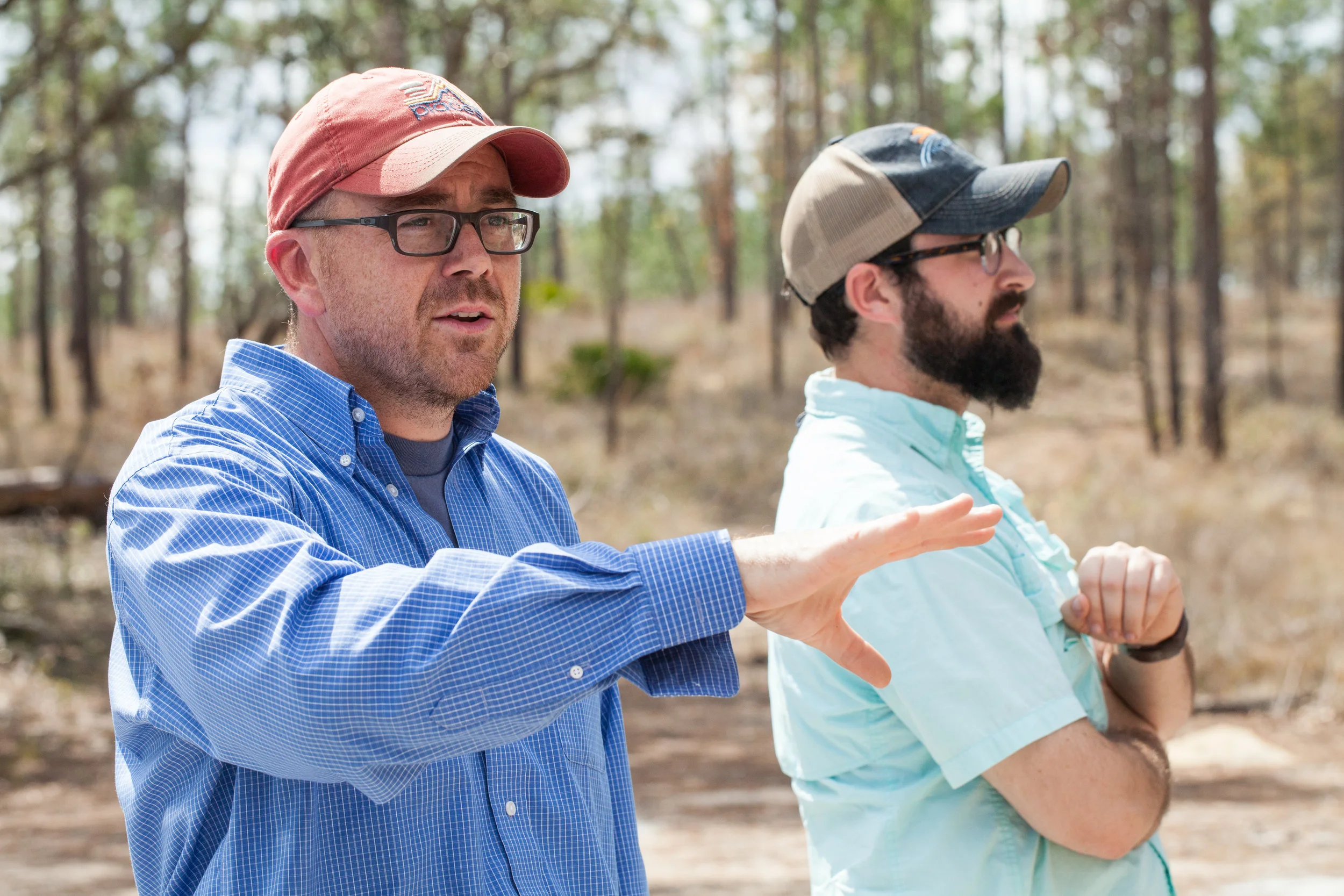 Two men outdoors in a wooded area, one is speaking and gesturing with his hand, the other is listening with arms crossed, both wearing casual shirts and caps.