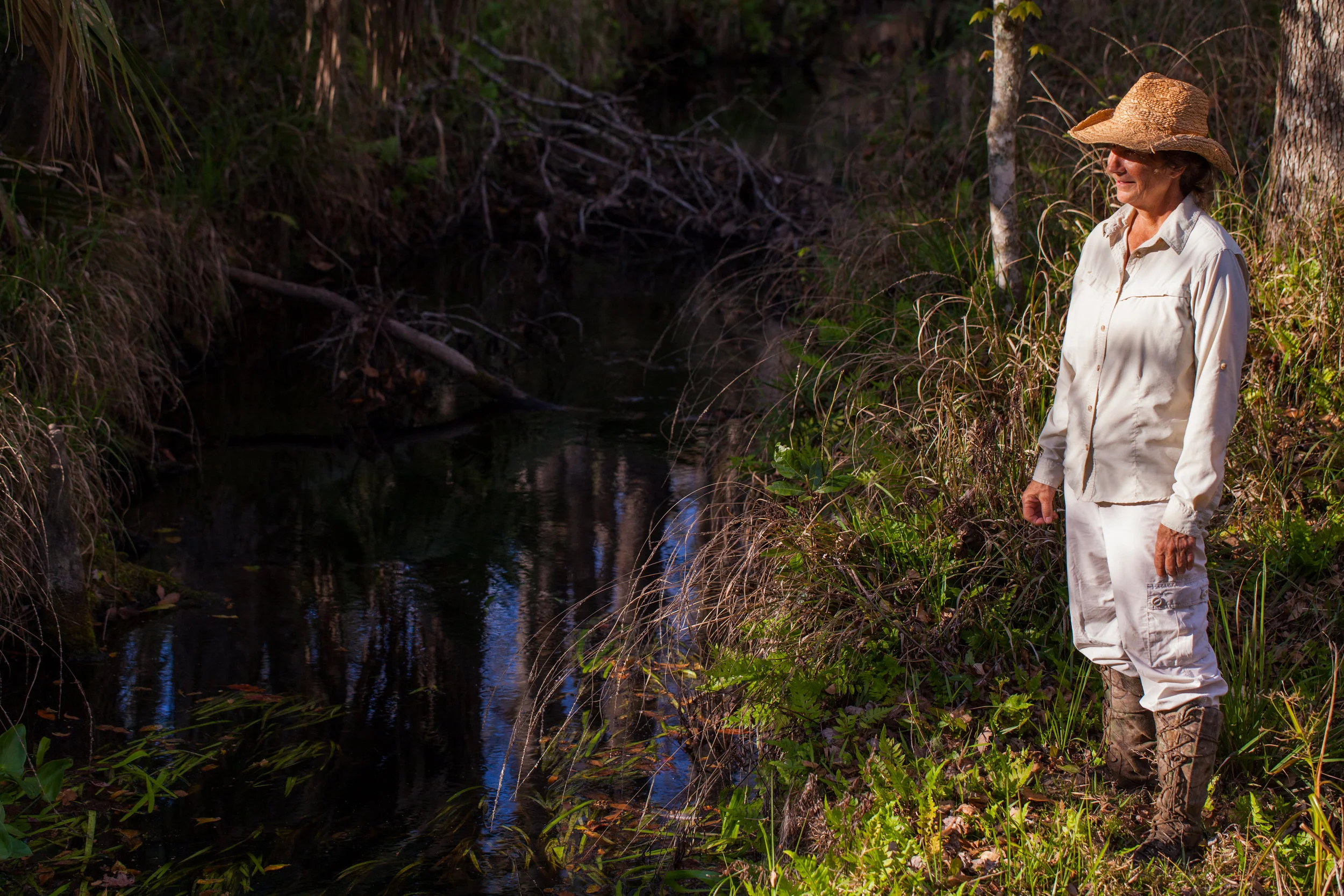 A woman wearing a wide-brimmed straw hat, a long-sleeved light-colored shirt, and white pants stands on the grassy bank of a small creek in a wooded area.