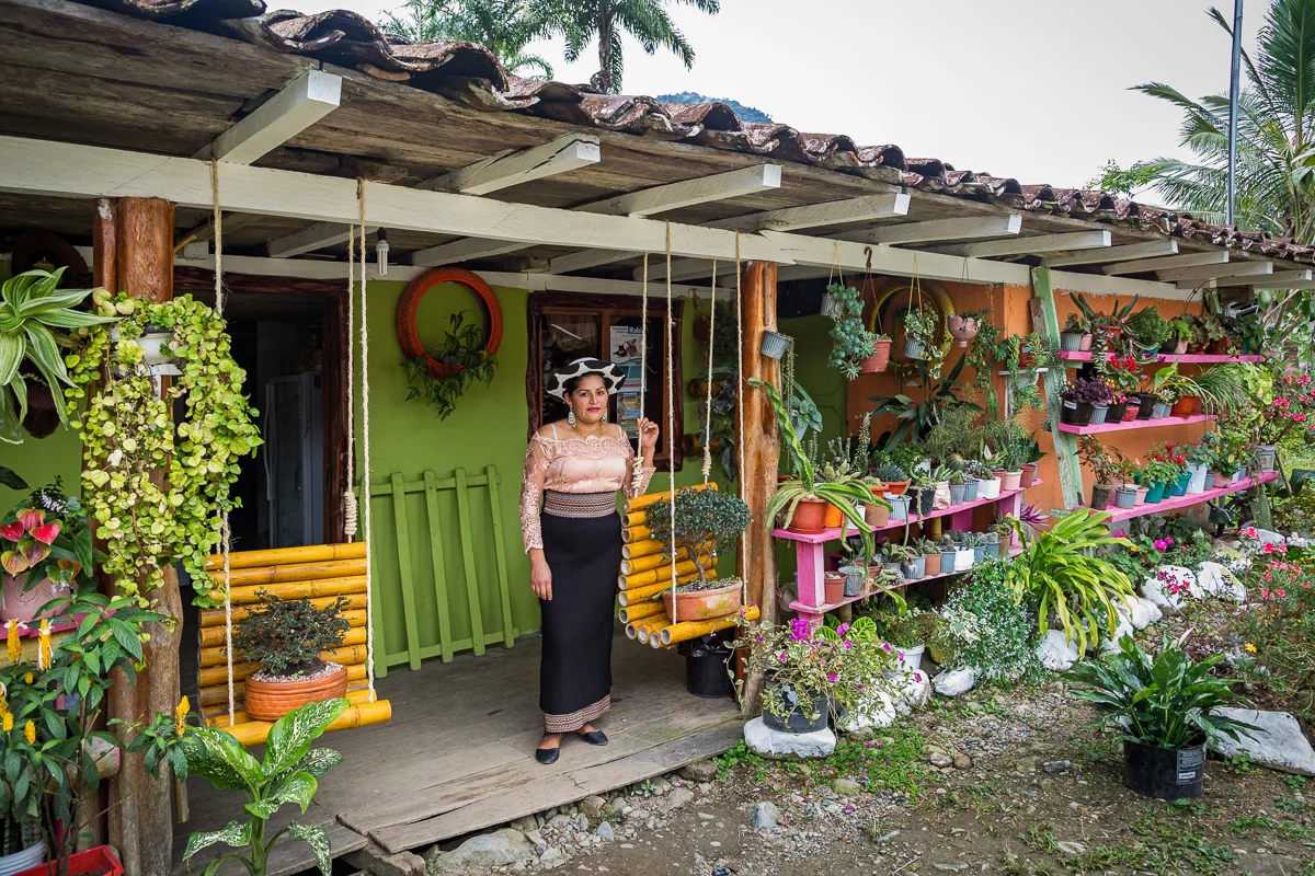 A woman standing on the porch of a colorful house surrounded by potted plants and flowers.