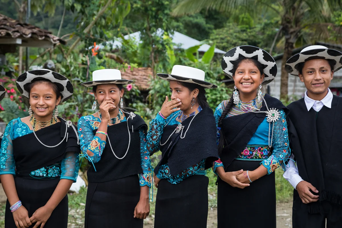 Group of five children dressed in traditional Saraguran attire in Ecuador, wearing black and blue embroidered clothing and black and white cow-patterned hats, standing outdoors surrounded by greenery and flowers.