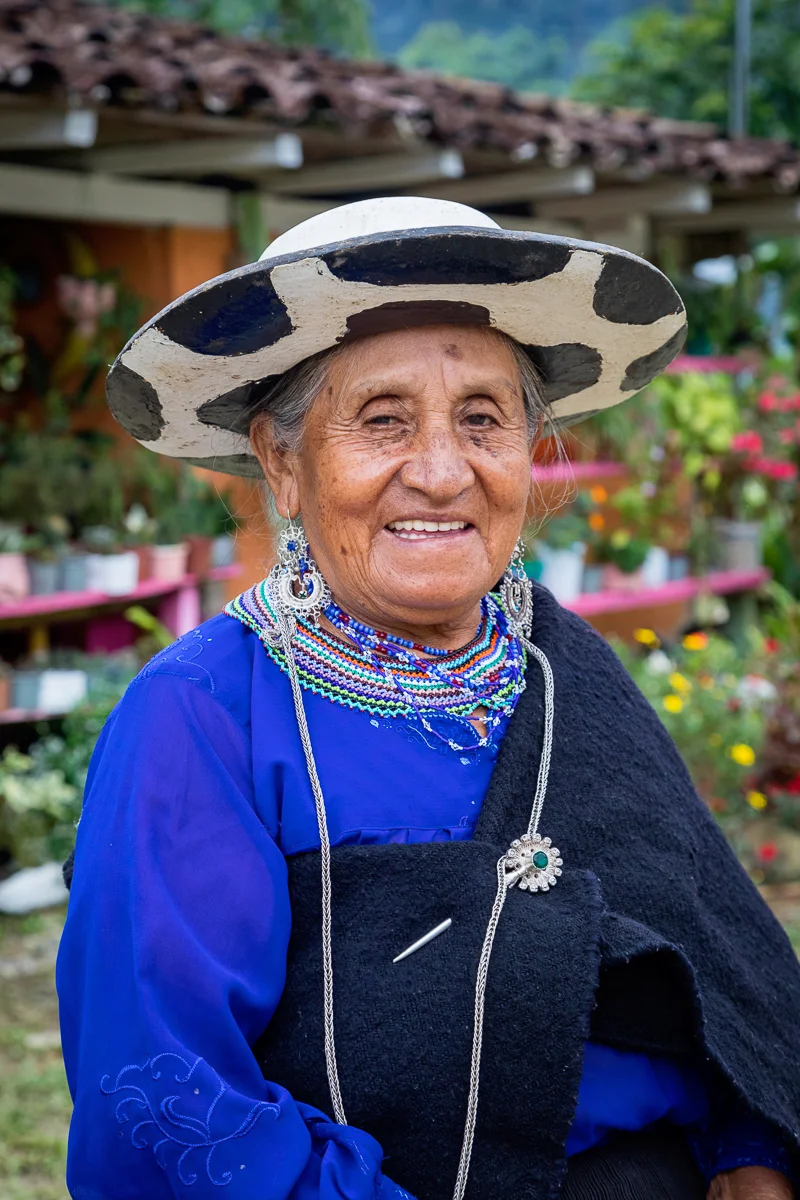 A smiling elderly woman in traditional clothing, wearing a large black and white hat, colorful beaded necklaces, earrings, and a black shawl, standing outdoors among greenery and flowers.