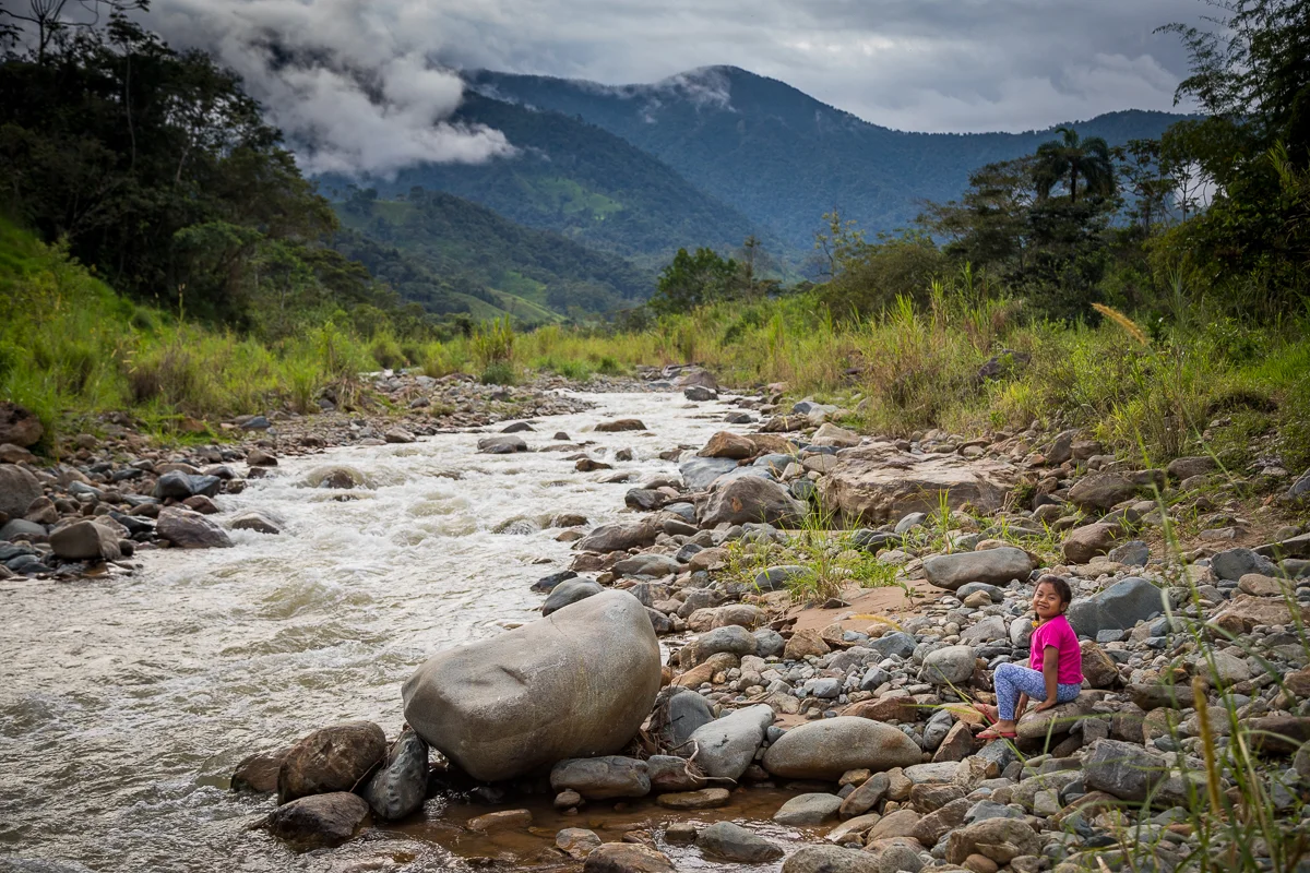 A young girl wearing a pink shirt and patterned pants is sitting on rocks by a rushing river, with lush green trees and mountains in the background and cloudy skies overhead.