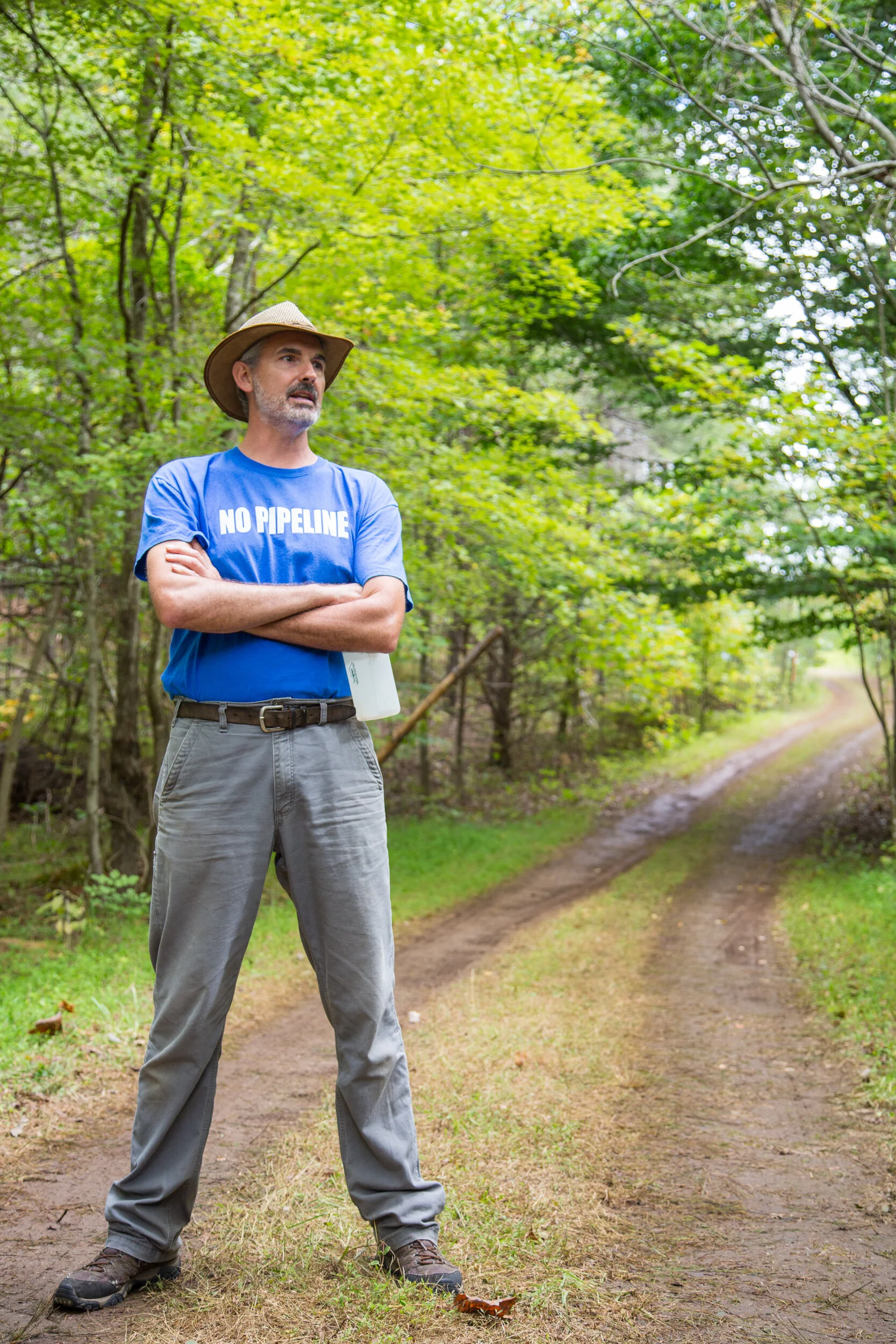 A man standing on a dirt path in a forest with trees and greenery, crossing his arms. He is wearing a straw hat, a blue t-shirt with the words 'NO PIPELINE', khaki pants, and hiking shoes.