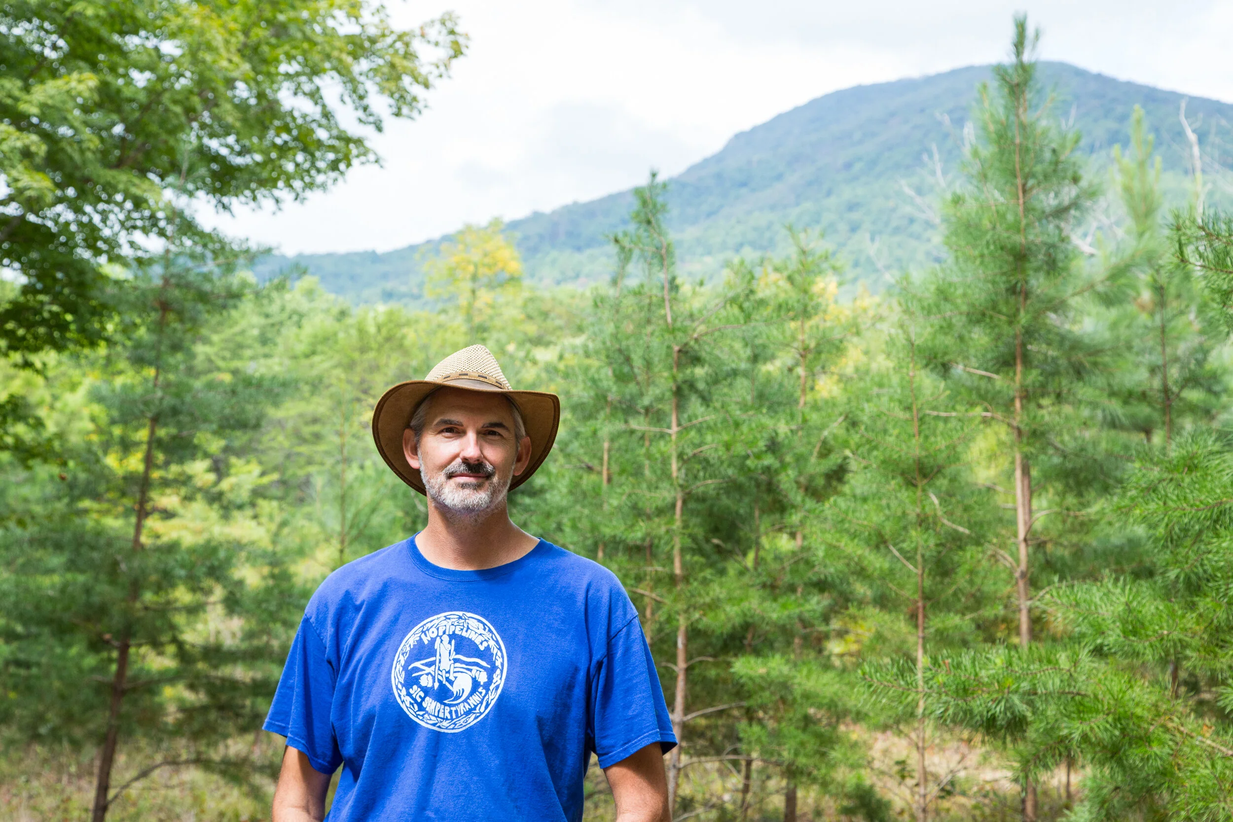A man with gray hair and beard wearing a wide-brimmed hat and a blue T-shirt with a circular logo stands outdoors in a green forest with mountains in the background.