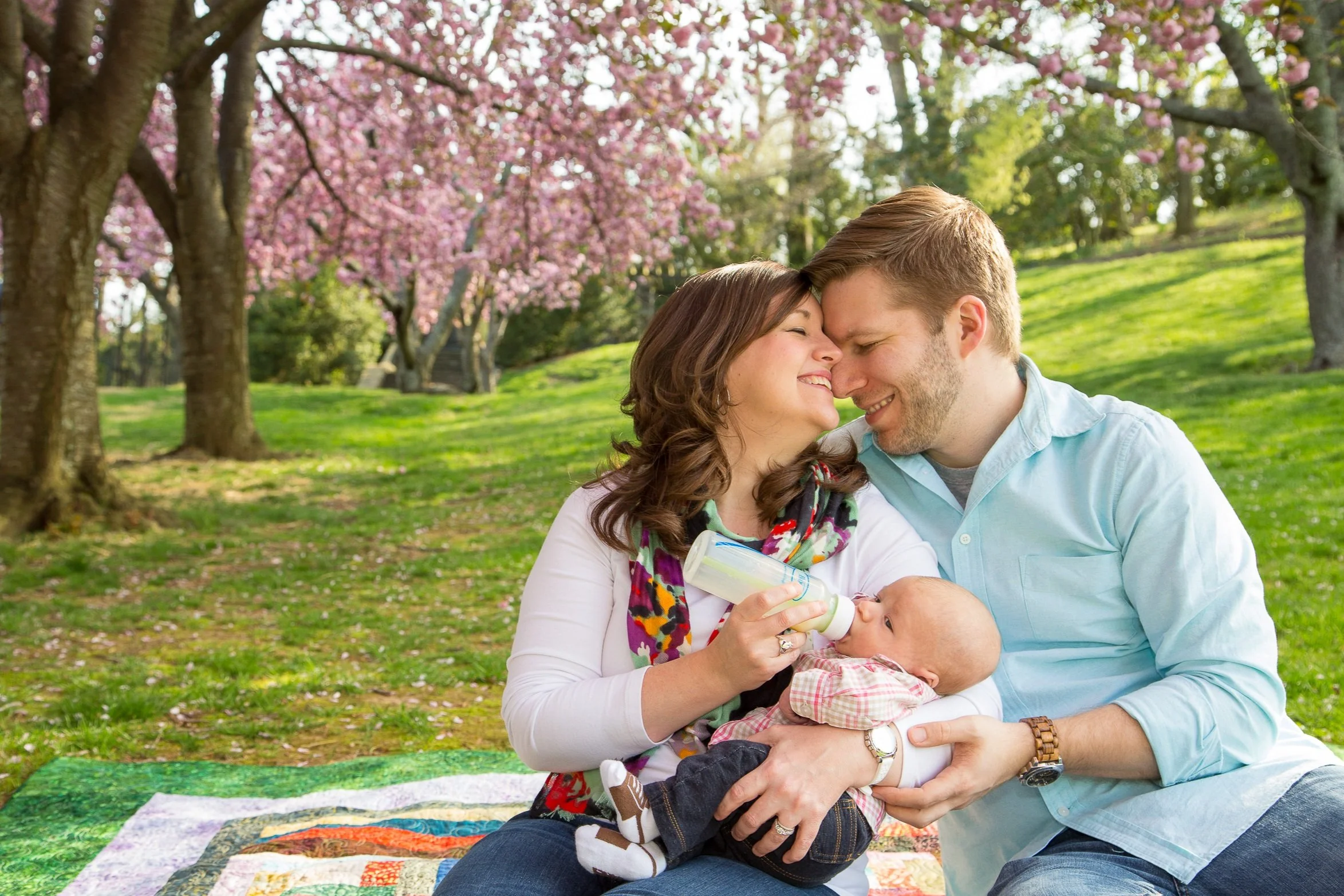 A happy family of three, feeding a baby with a bottle, sitting on a blanket in a park with pink cherry blossom trees in the background.