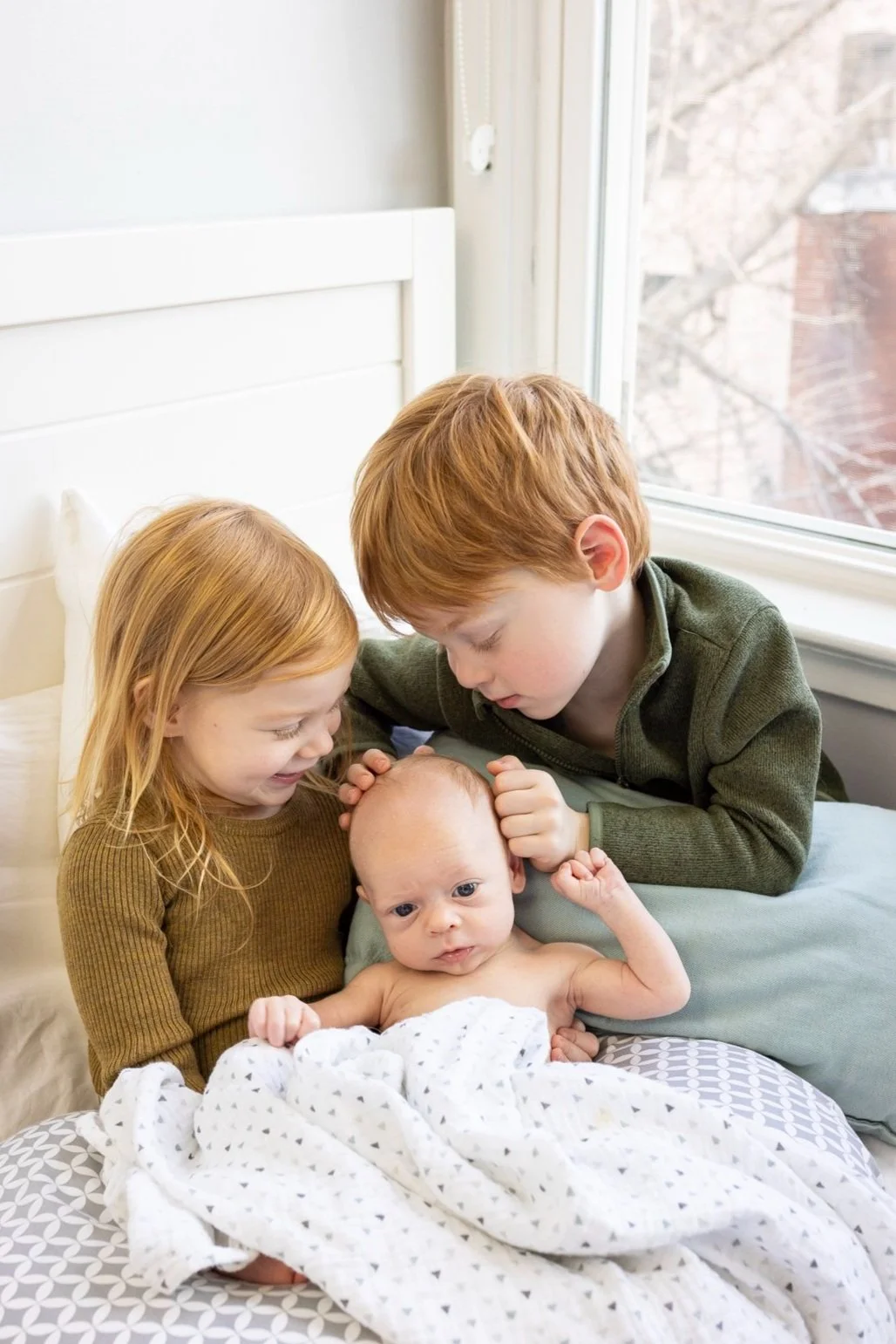 Two older children, a girl and a boy, are sitting on a bed with a newborn baby. The girl and boy are gently touching the baby's head, looking at the baby with affection. The scene is bright, next to a large window.