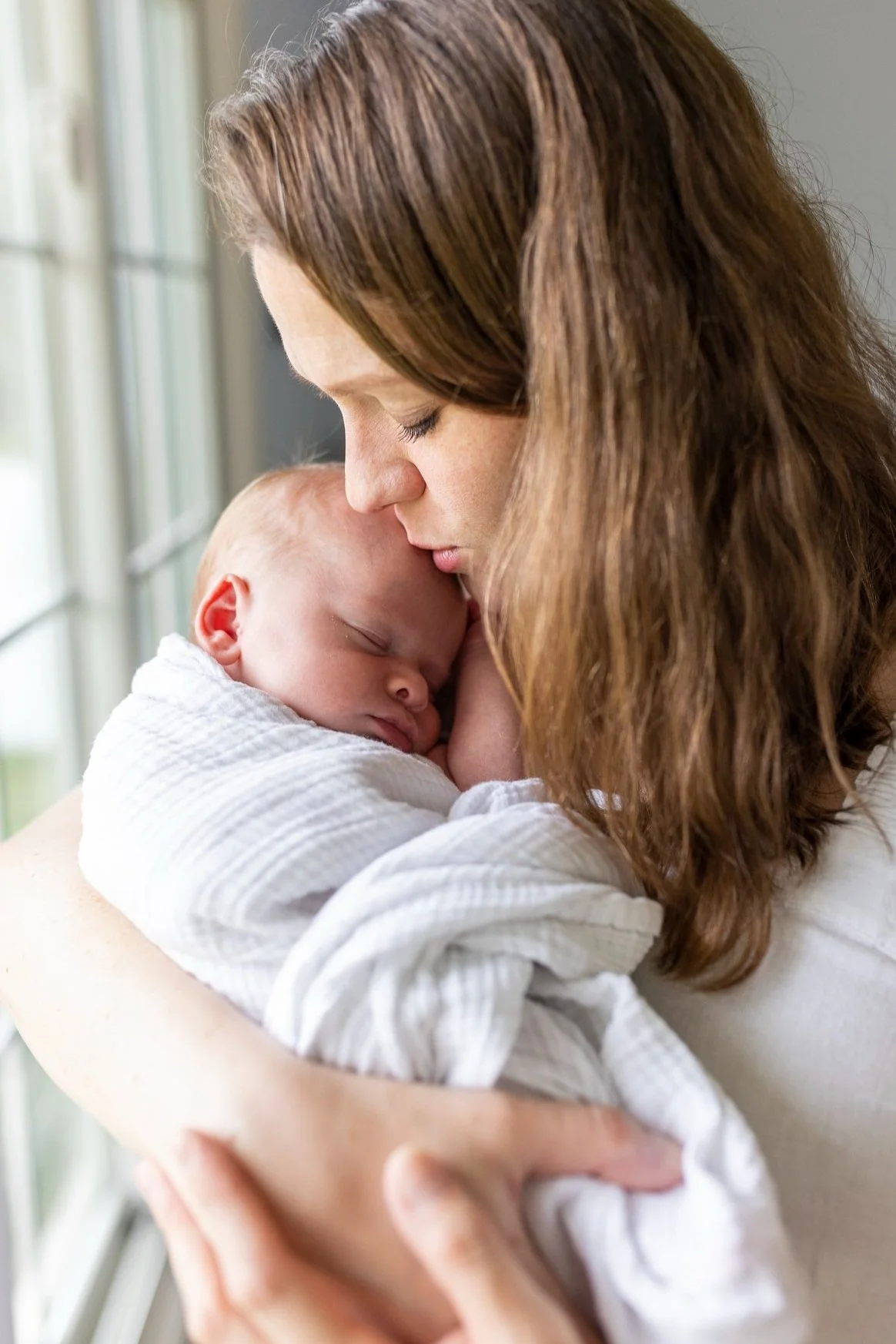 A mother holding her sleeping baby close, with her forehead touching the baby's head, near a window with natural light.