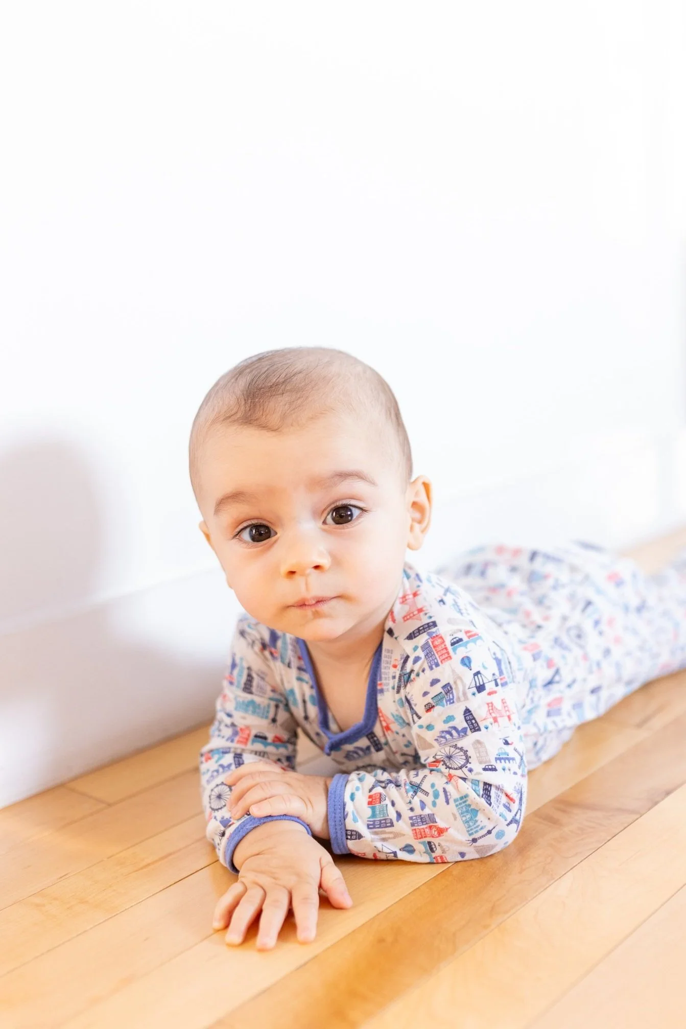 A baby lying on a wooden floor, wearing a colorful, patterned pajamas with a cityscape design, looking directly at the camera with a curious expression.