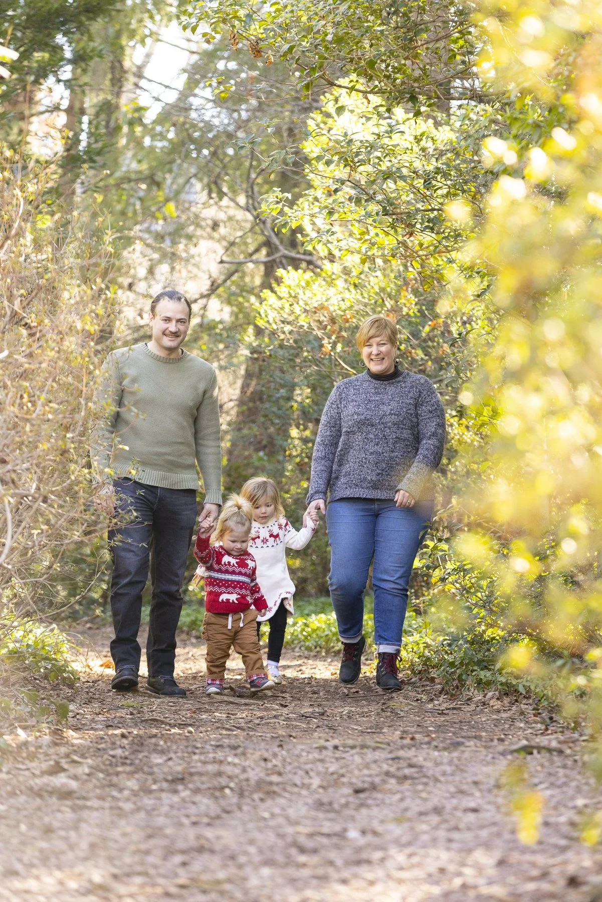 A family of four walking on a wooded trail in autumn, holding hands and smiling.