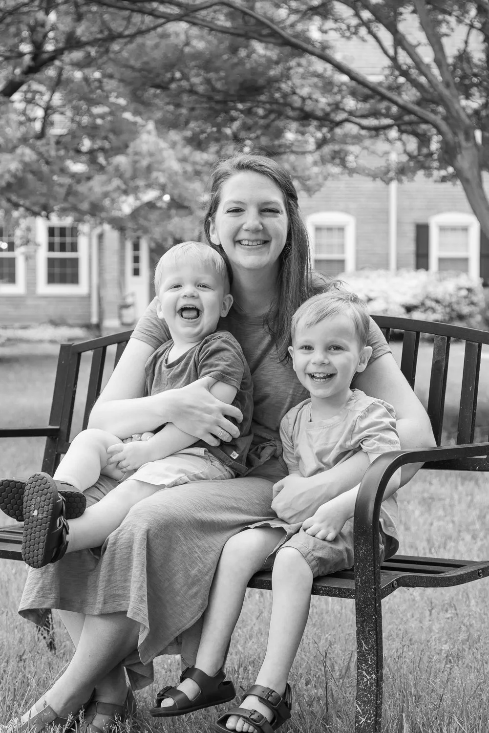 A woman sitting on a park bench with two young children, all smiling and laughing. The background shows trees and a house.