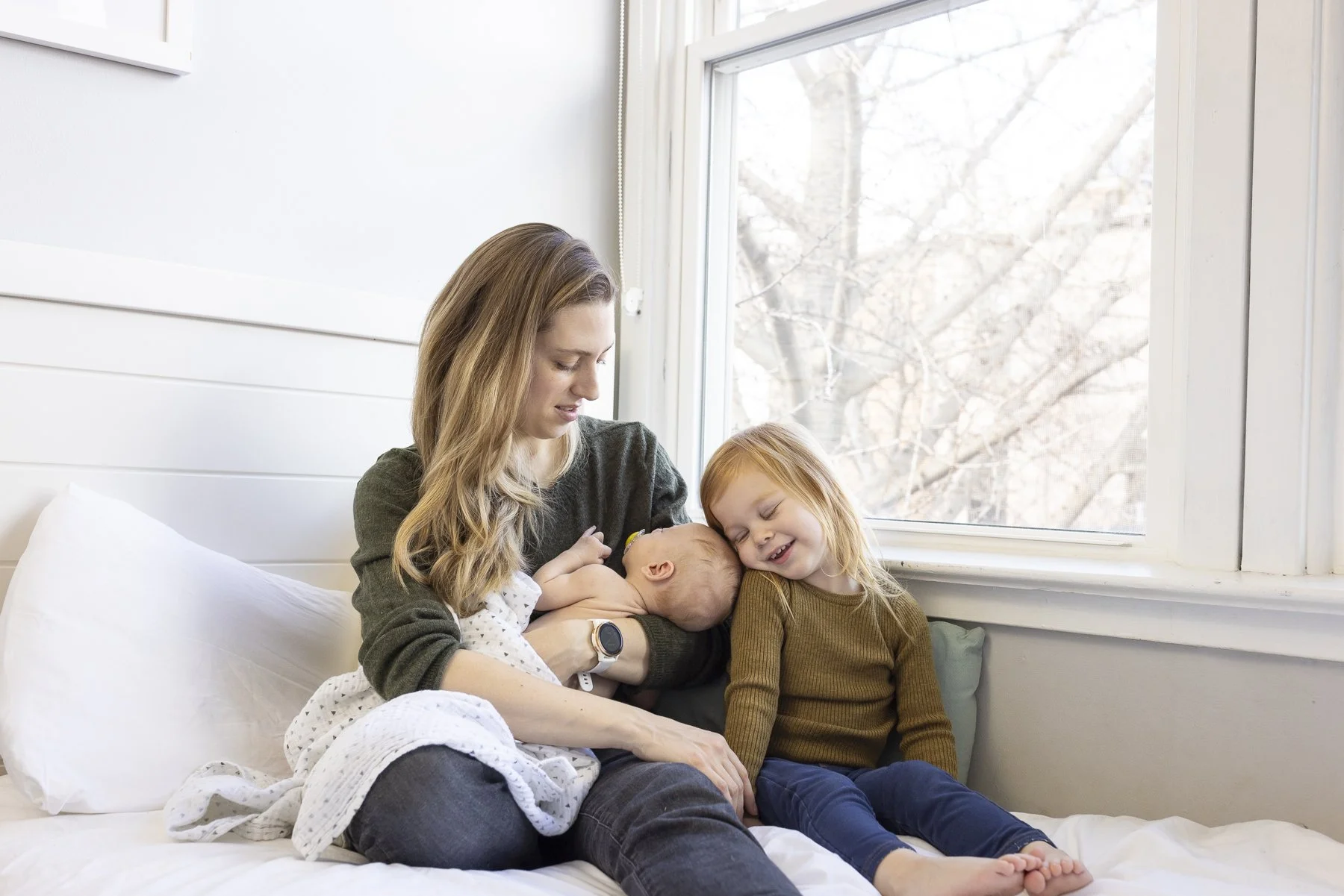 Newborn lifestyle portrait on a bed with mom and older sibling giving cuddles.