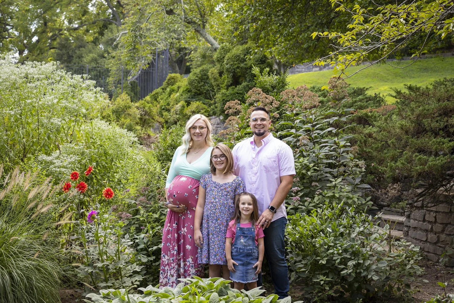 A family of four standing in a lush garden with green trees and bushes. The mother, with blonde hair and glasses, is pregnant and wearing a pastel blue top and pink floral skirt. The father, with dark hair and glasses, is wearing a light pink shirt a