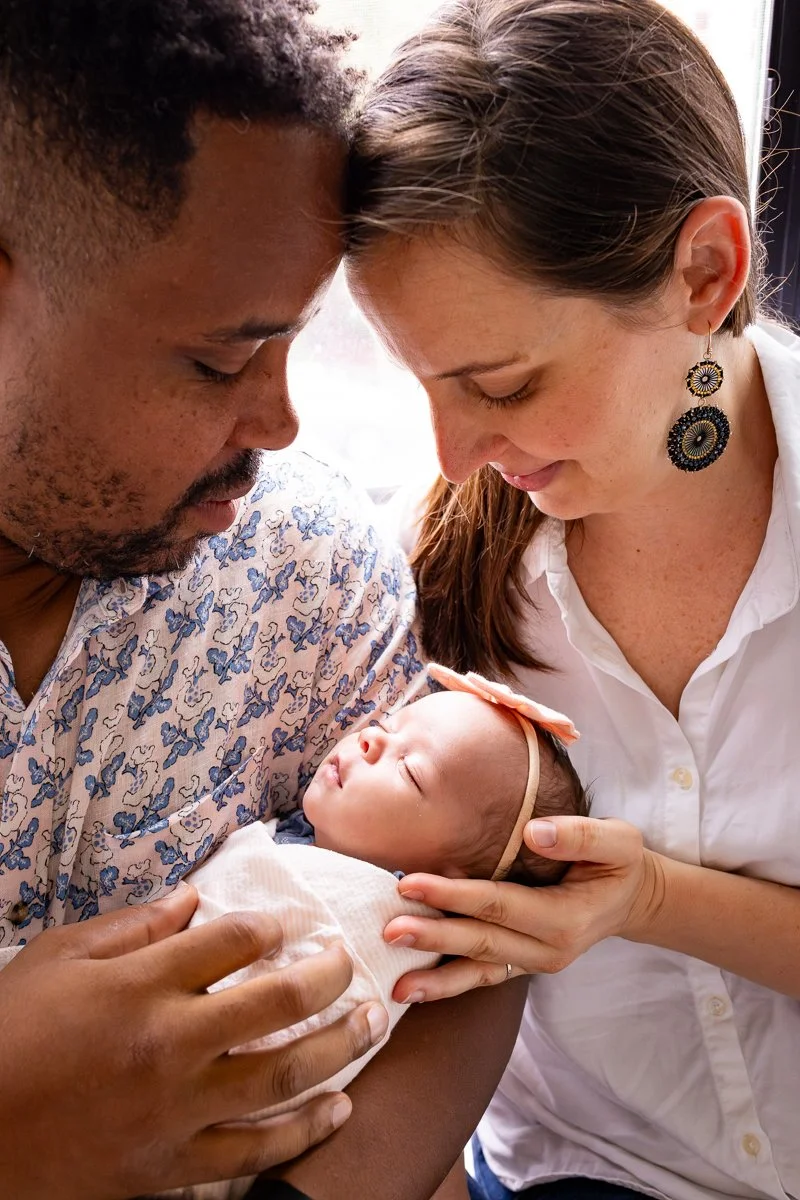A man and a woman gently holding and touching a sleeping baby girl with a pink headband and bow, in a close and tender moment.
