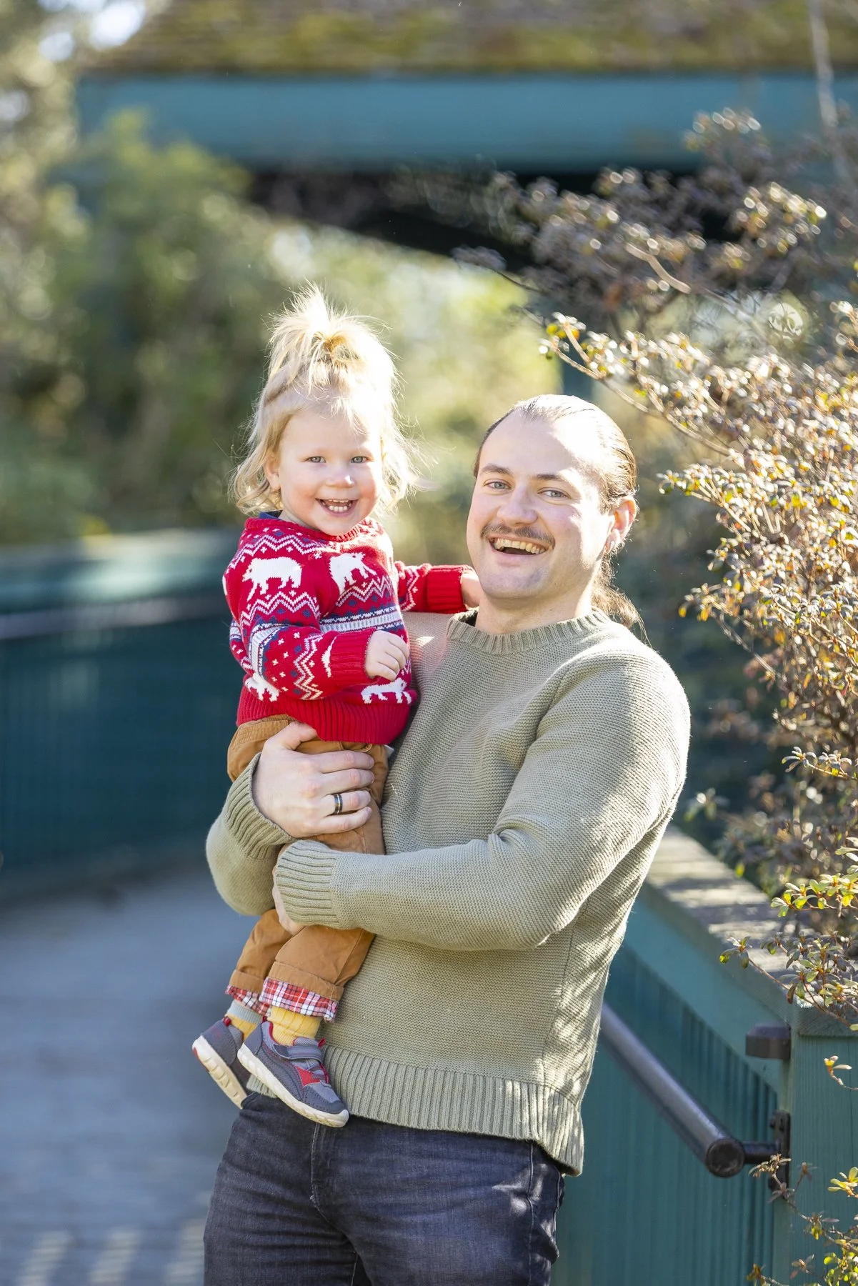 A man holding a young girl, both smiling, outdoors with trees and a blue bridge in the background.