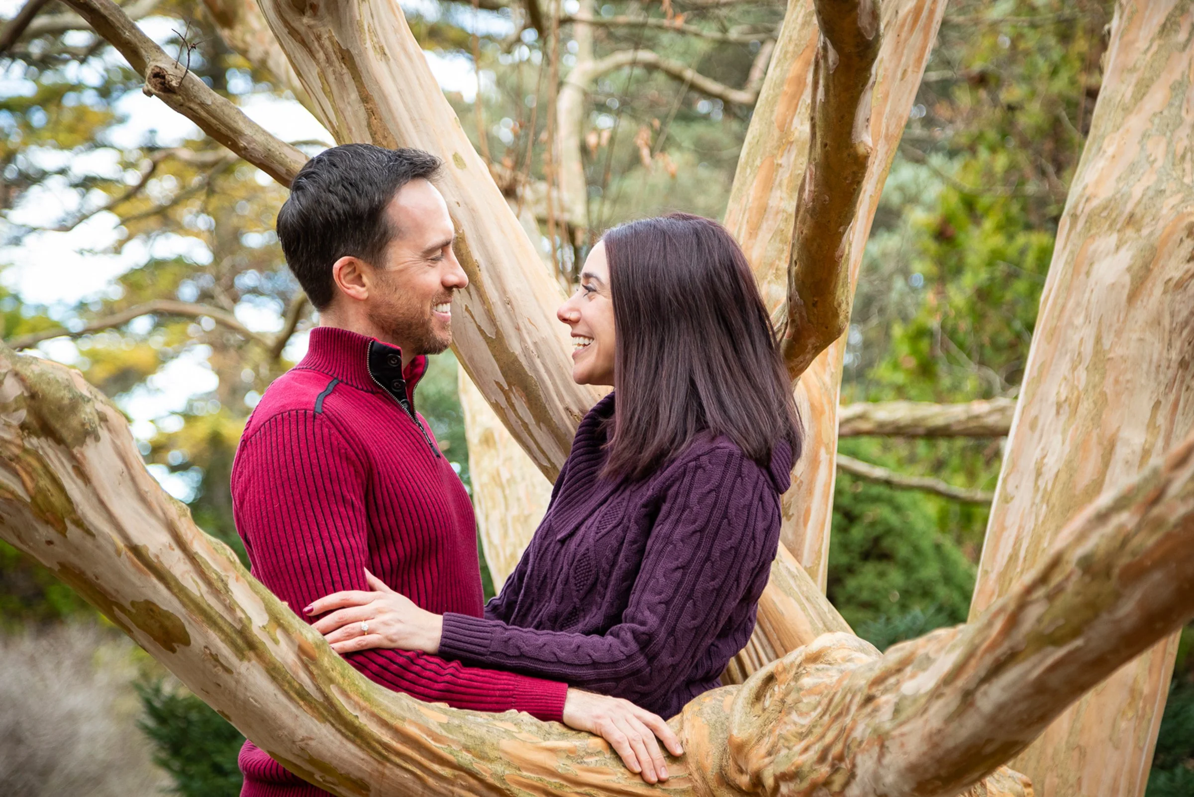A couple standing close to each other in a tree, smiling warmly and gazing into each other's eyes, outdoors surrounded by greenery.