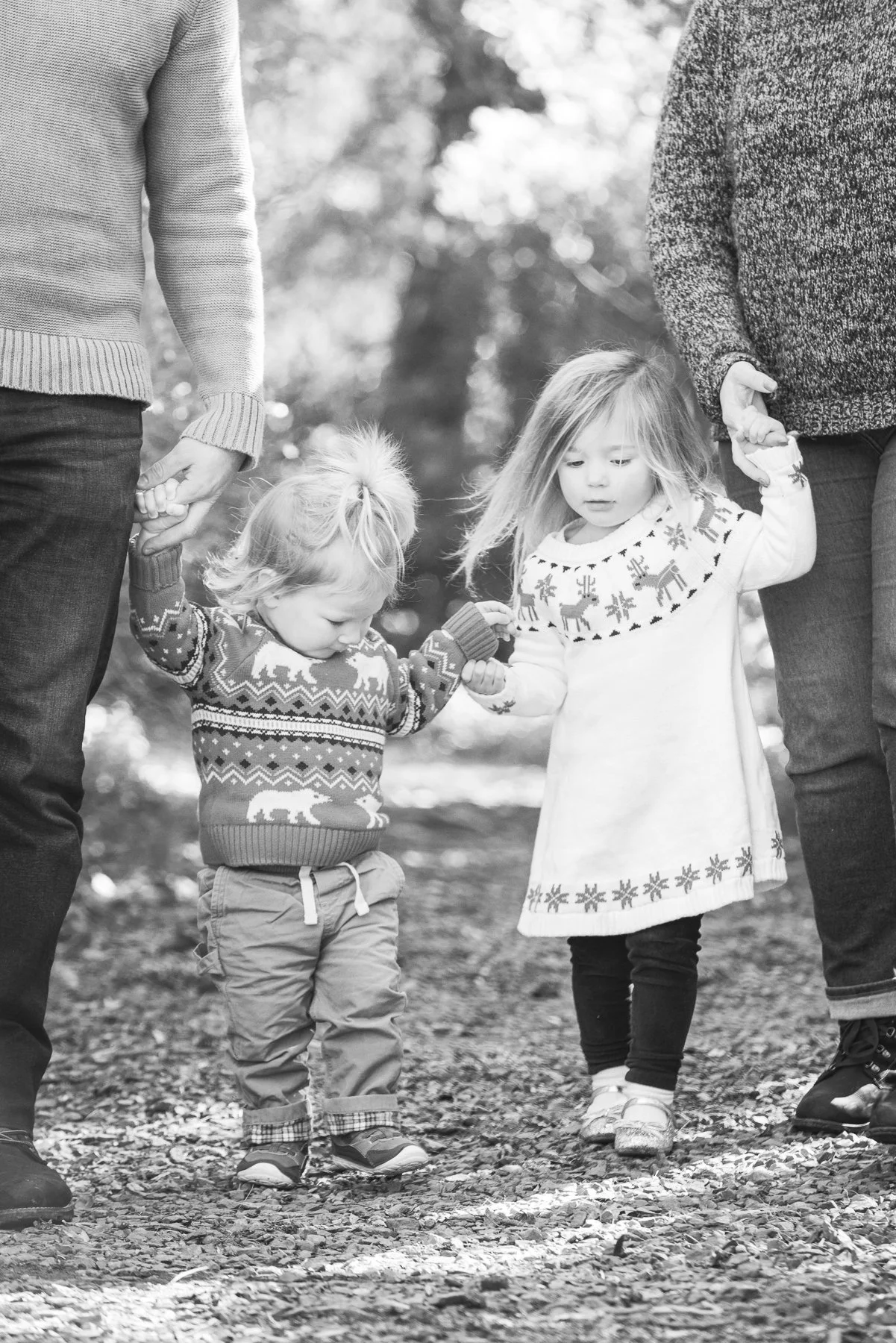 Two young children holding hands with adults on a nature trail, wearing Christmas sweaters, in a black-and-white photo.