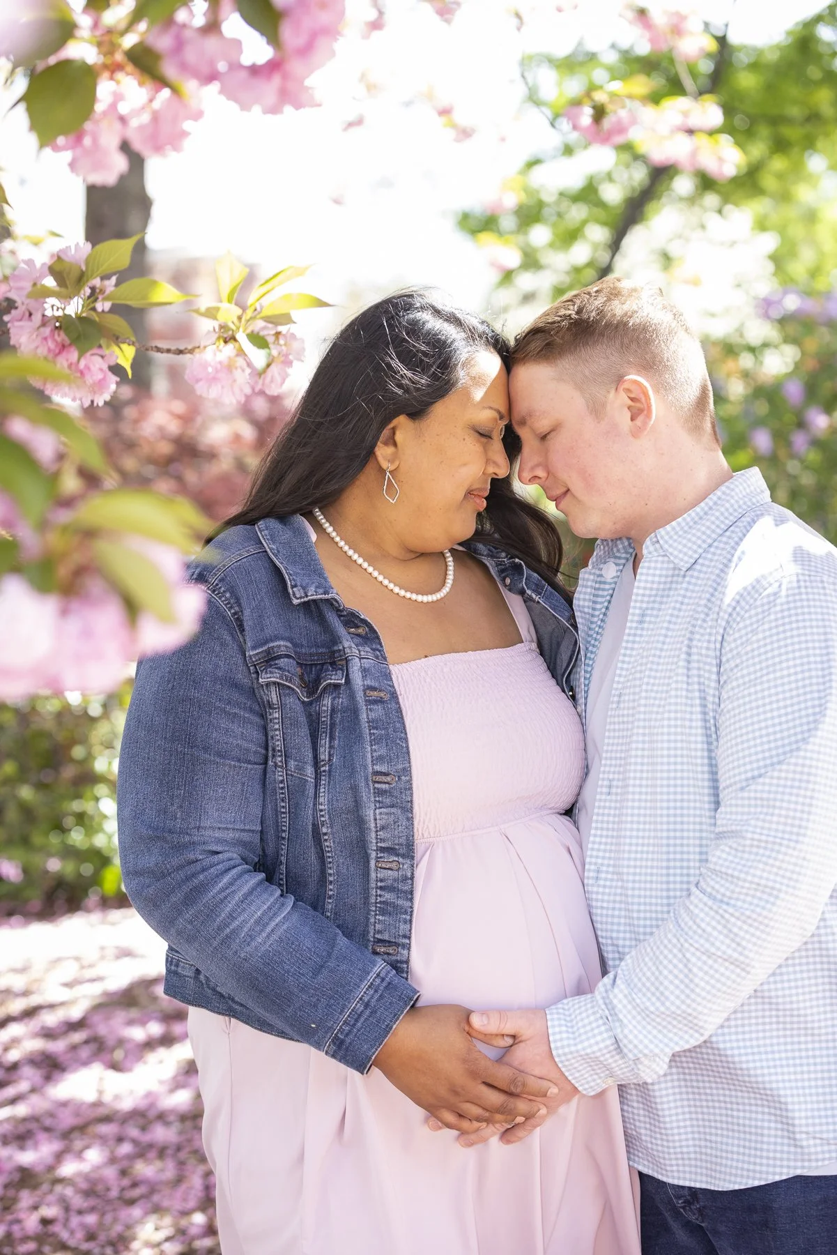 An expecting mother and her partner holding hands, with their foreheads touching in a gentle hug, amid blooming pink and purple flowers in a garden.