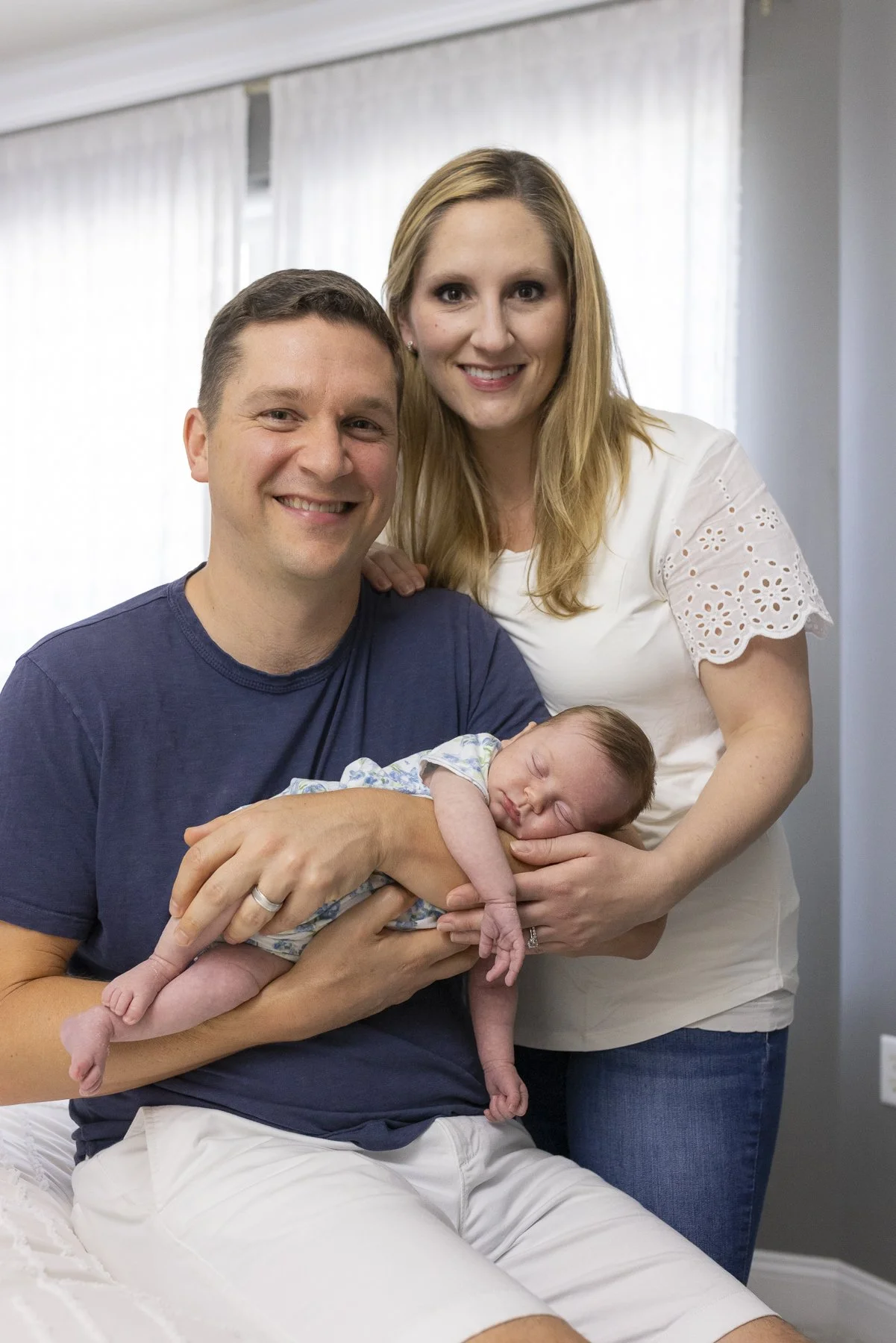 A smiling couple holding a sleeping baby in a room with white curtains