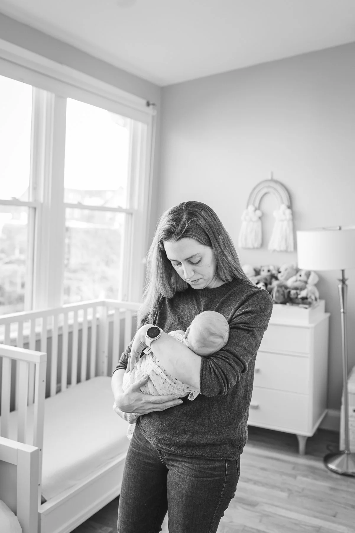 A woman holding a newborn baby in a nursery with large windows, a white crib, and a dresser with flowers in the background.