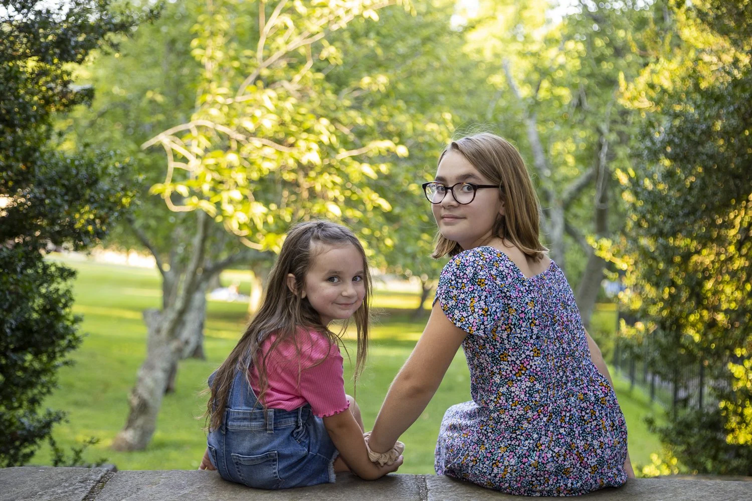 Two young girls sitting on a stone wall outdoors, holding hands, with trees and greenery in the background during the day.
