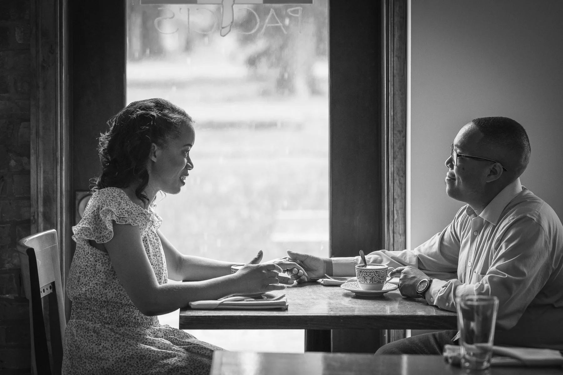 A woman and a man are sitting across from each other at a table in a cafe, holding hands and gazing into each other's eyes.
