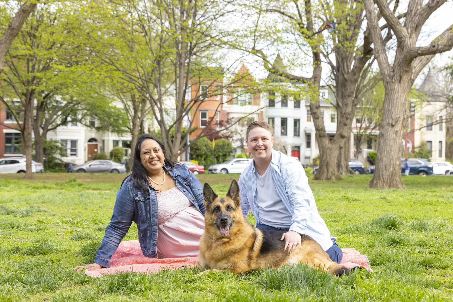 A diverse couple with a German Shepherd dog sitting on a blanket on a grassy park with trees and houses in the background.