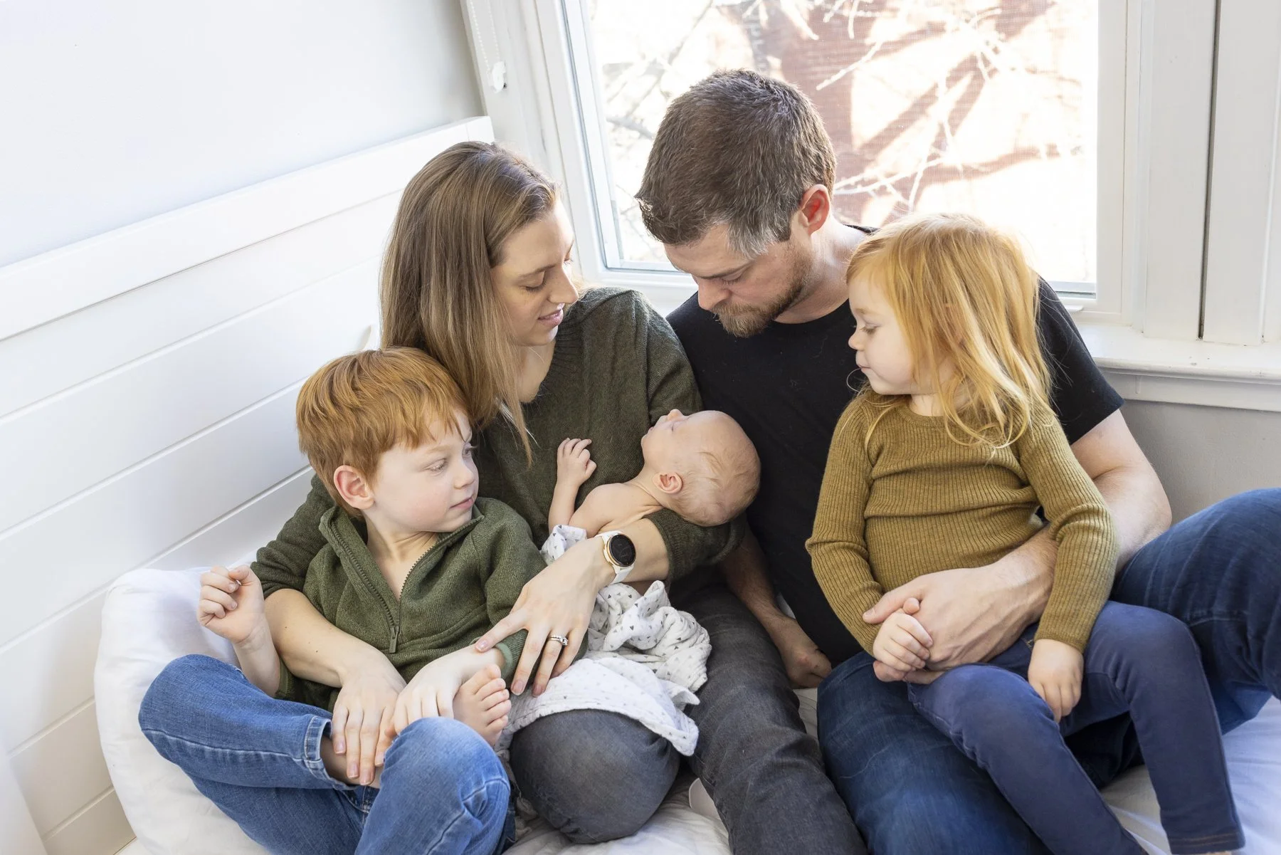 Family of five sitting closely on a bed, breastfeeding and cuddling, by a window. Two young children, a boy and a girl, are looking at a newborn baby girl, all enjoying a tender moment.