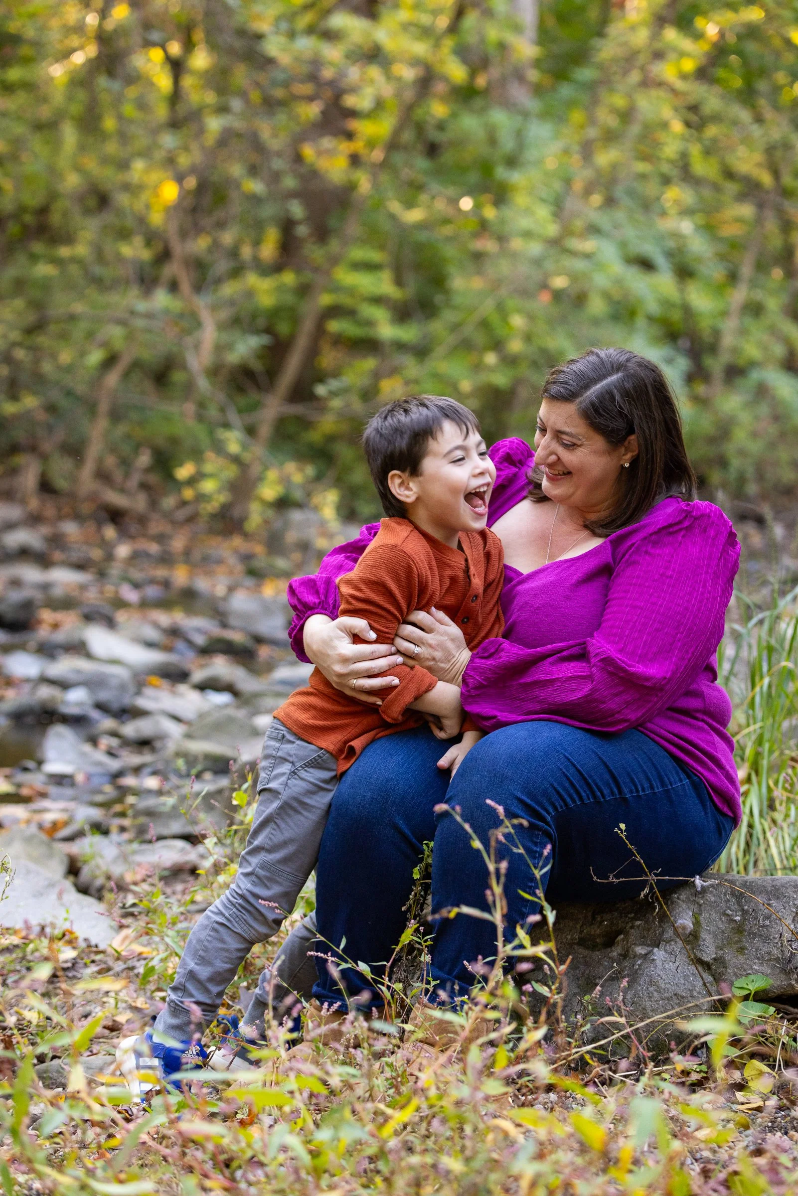 A woman and a young boy sharing a joyful moment outdoors in a forest, with the woman sitting on a rock and the boy laughing, surrounded by autumn foliage.