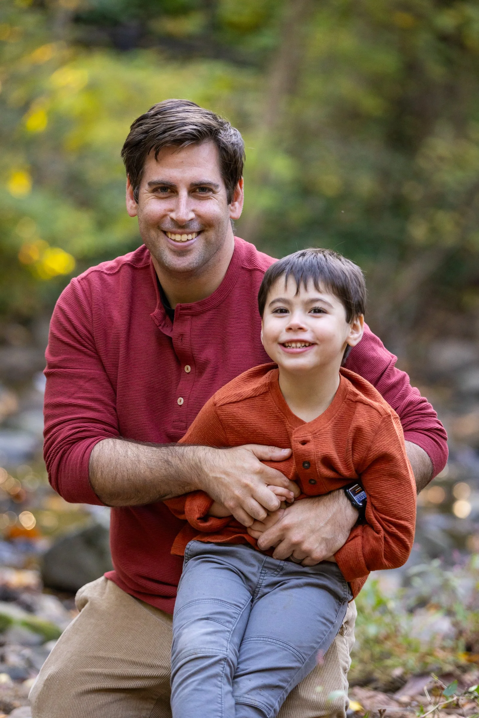 A man and a young boy smiling outdoors in a natural setting, standing near a stream with trees in the background.