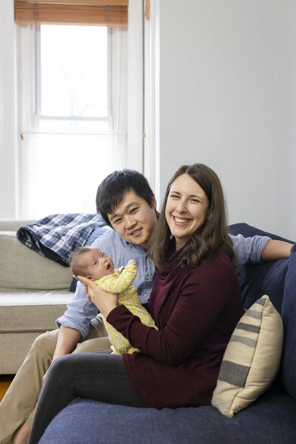 A happy family of three, including a woman holding a newborn baby and a man, sitting on a blue sofa in a cozy, well-lit living room.