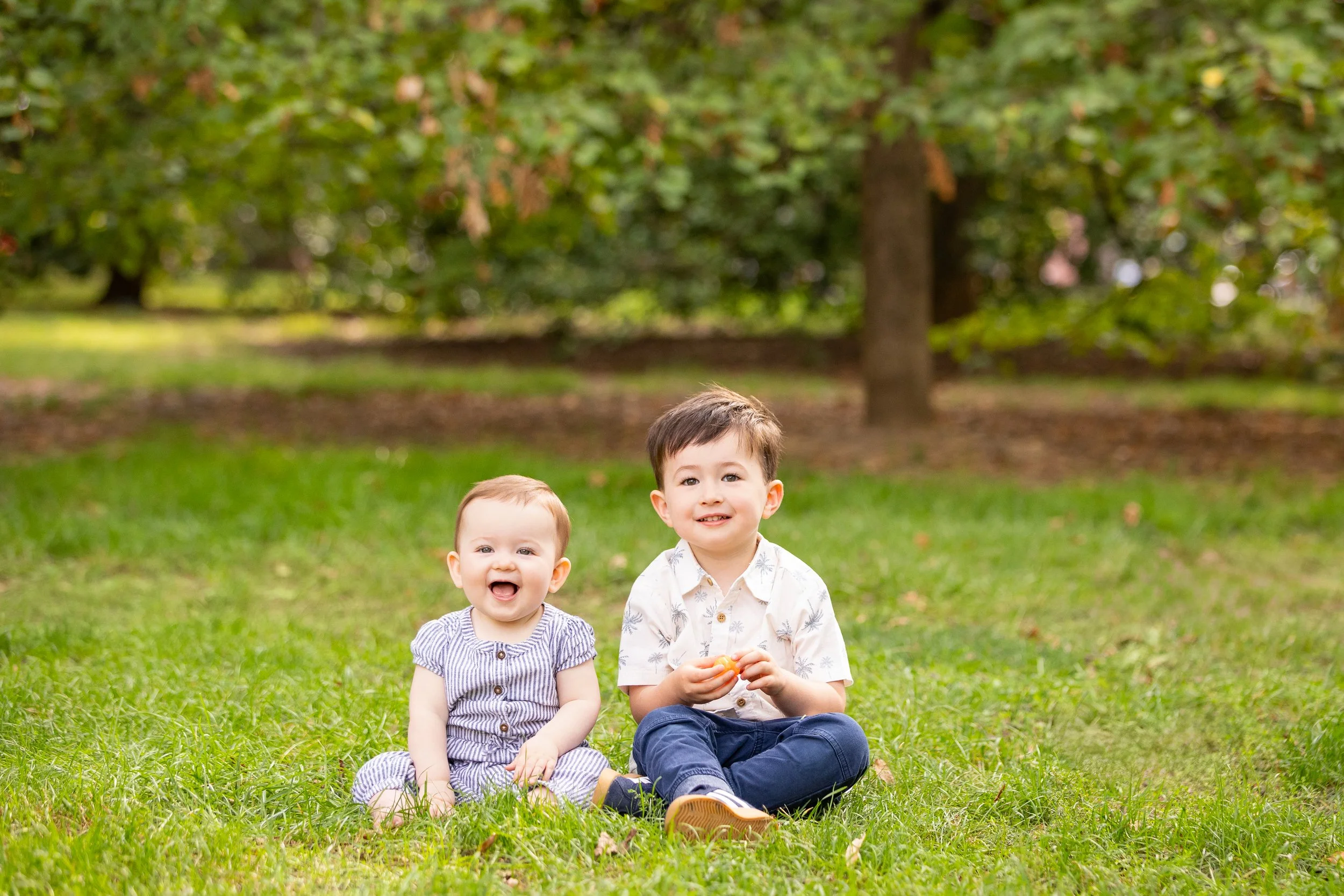 Two young children, a girl and a boy, sitting on green grass in a park with trees in the background, smiling and enjoying a day outdoors.