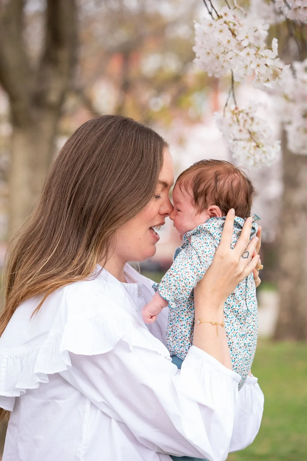 A woman holding a newborn baby outdoors near blooming cherry blossom trees, both with noses touching.