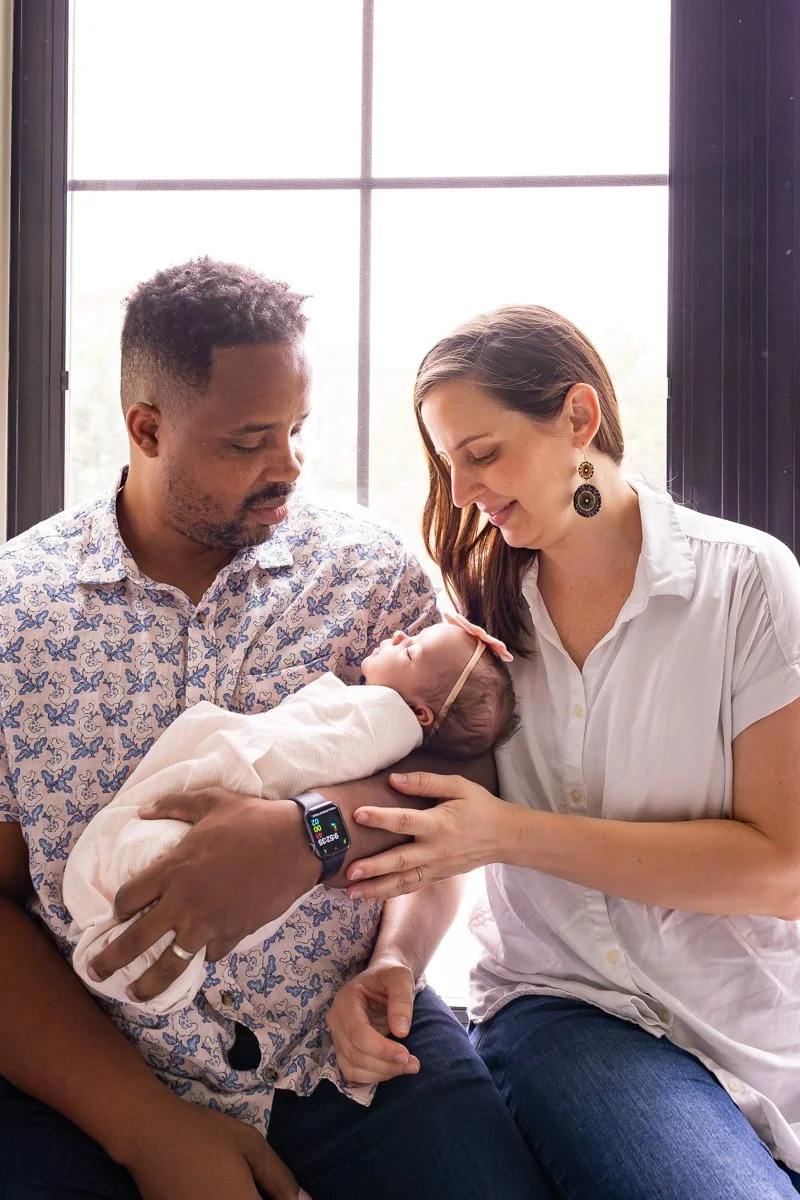 A man and woman holding a newborn baby together, looking at the baby with affection, inside a room with large window behind them.
