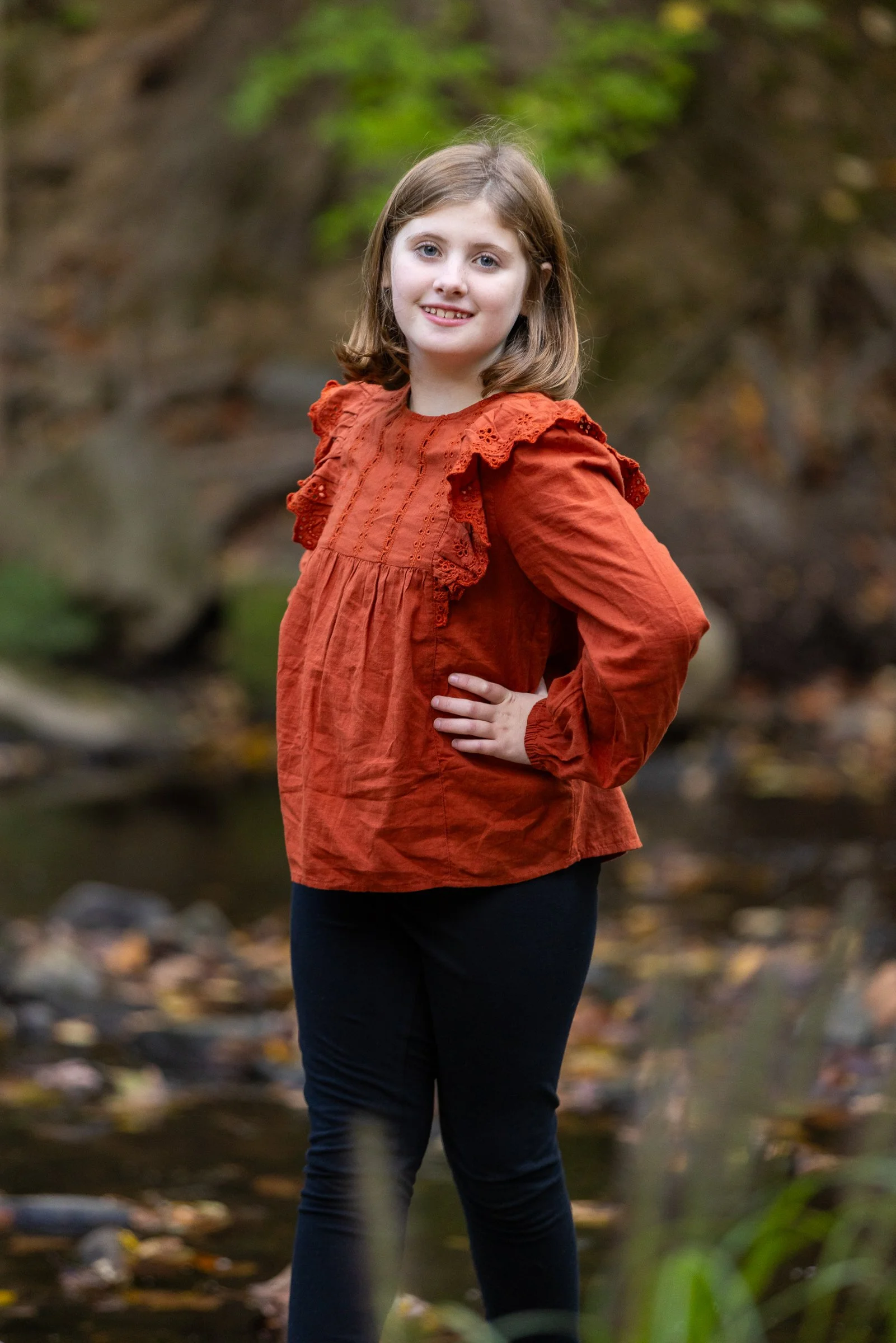 A young girl with shoulder-length light brown hair, smiling with her hand on her hip, stands outdoors near a stream in a wooded area during autumn, wearing an orange blouse and dark pants.
