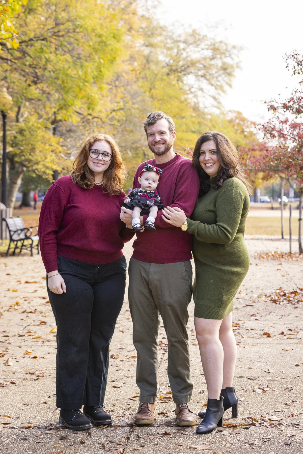 A family of four, including a woman, a man, a teenage girl, and a baby girl, standing outdoors in a park with fall foliage. The family is smiling, and the baby girl is being held by the man.