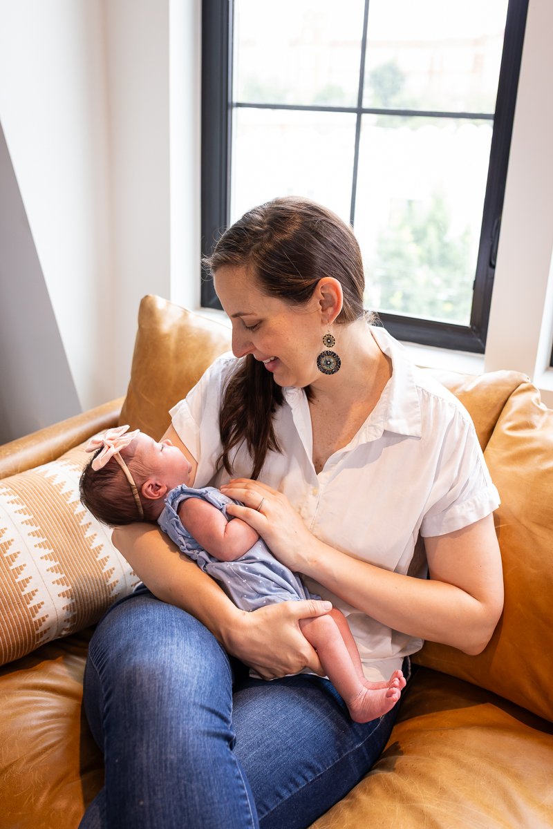 A woman sitting on a tan leather couch, holding a newborn baby girl in her arms, looking at the baby adoringly with a smile, inside a room with a large window behind her.