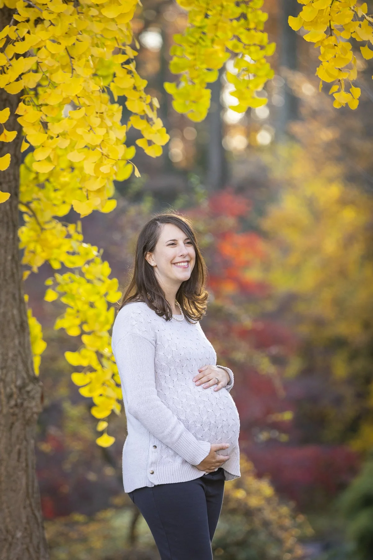Pregnant woman posing outdoors among autumn trees, smiling and holding her belly with both hands.