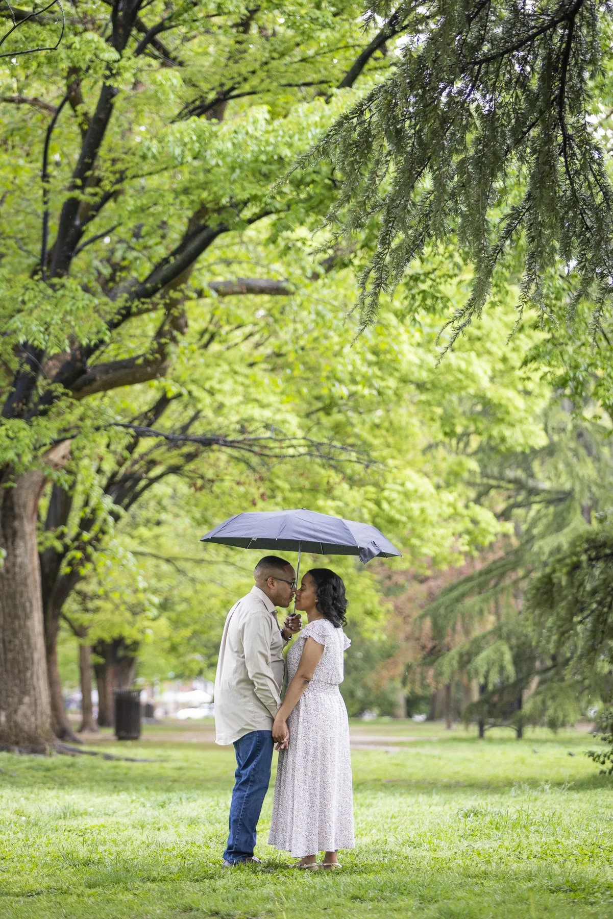 A couple standing close together under a black umbrella in a lush, green park, about to kiss.