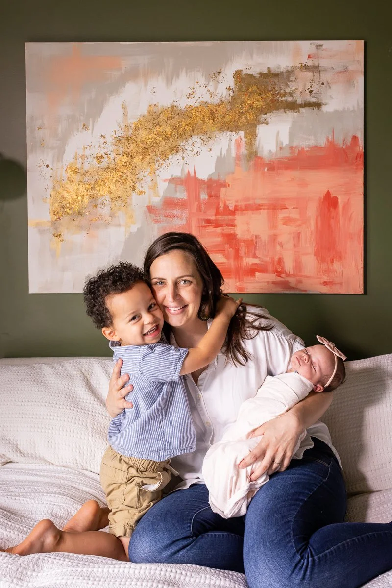 A woman with two young children, one girl and one boy, sitting on a cream-colored couch in front of an abstract painting with shades of pink, gray, white, and gold accents. The woman is smiling and holding the girl, who is hugging her, and the girl i