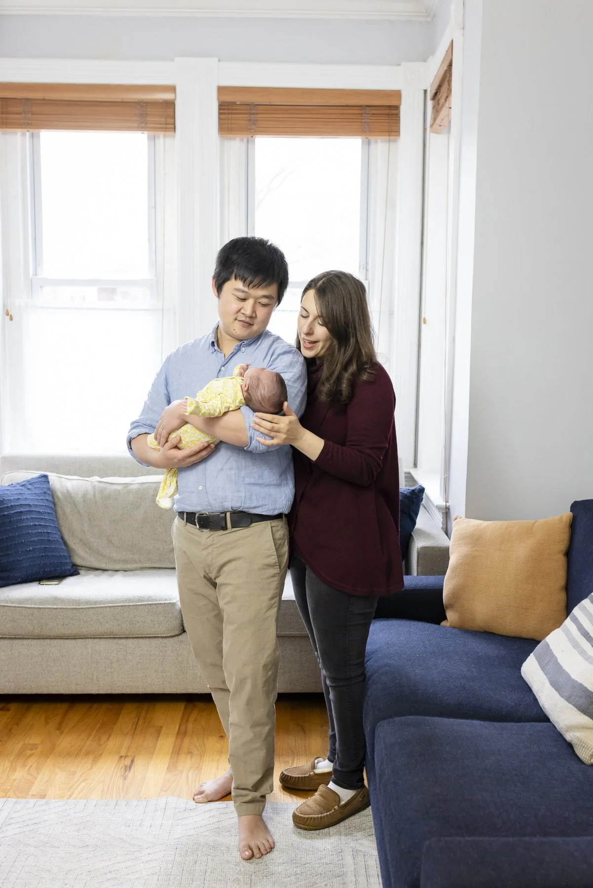 A young couple with a newborn baby in a living room with large windows and a beige and blue couch.