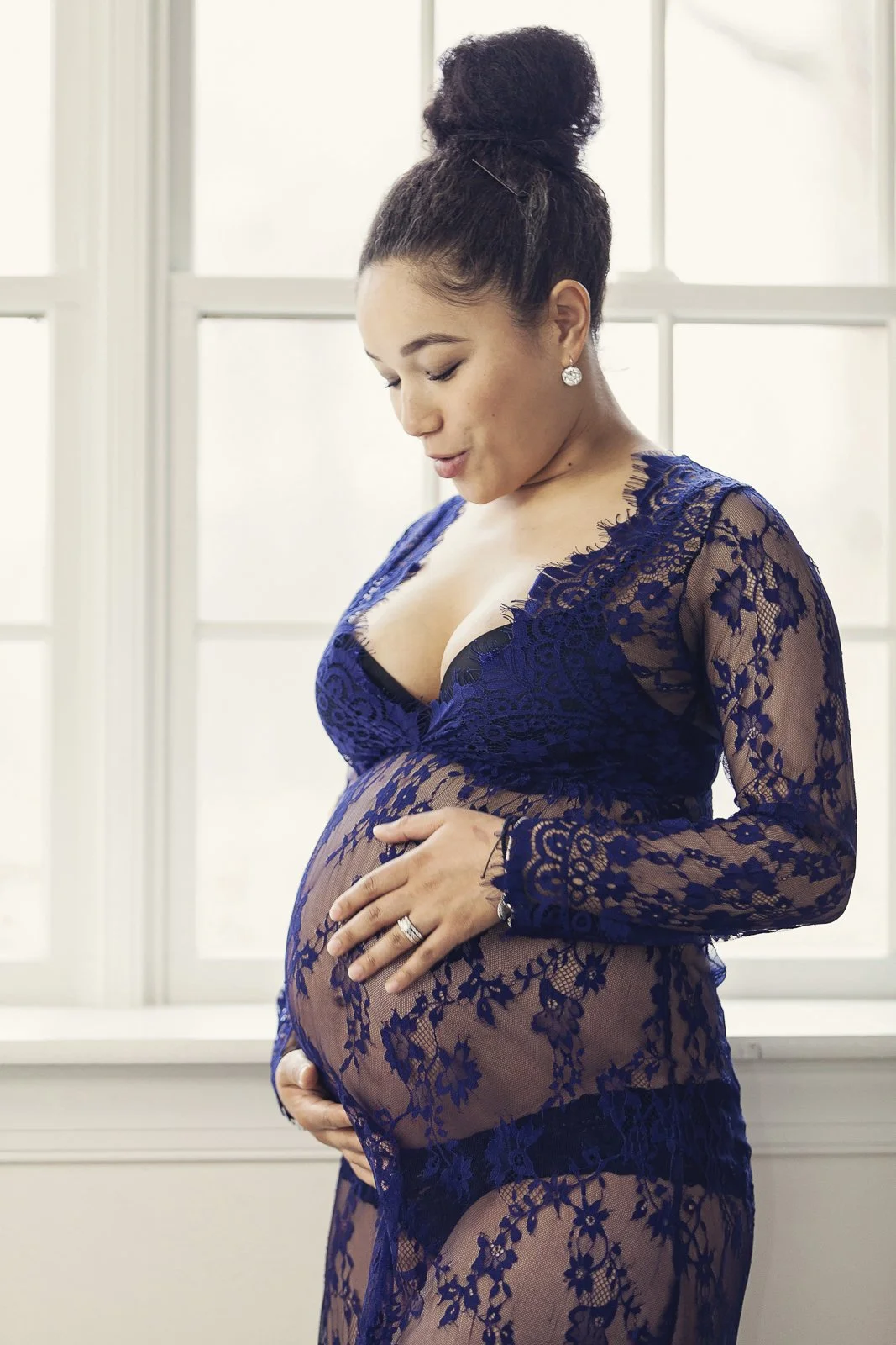 A pregnant woman in a blue lace dress, standing in front of a window, gently cradling her baby bump with both hands, looking down at her belly.